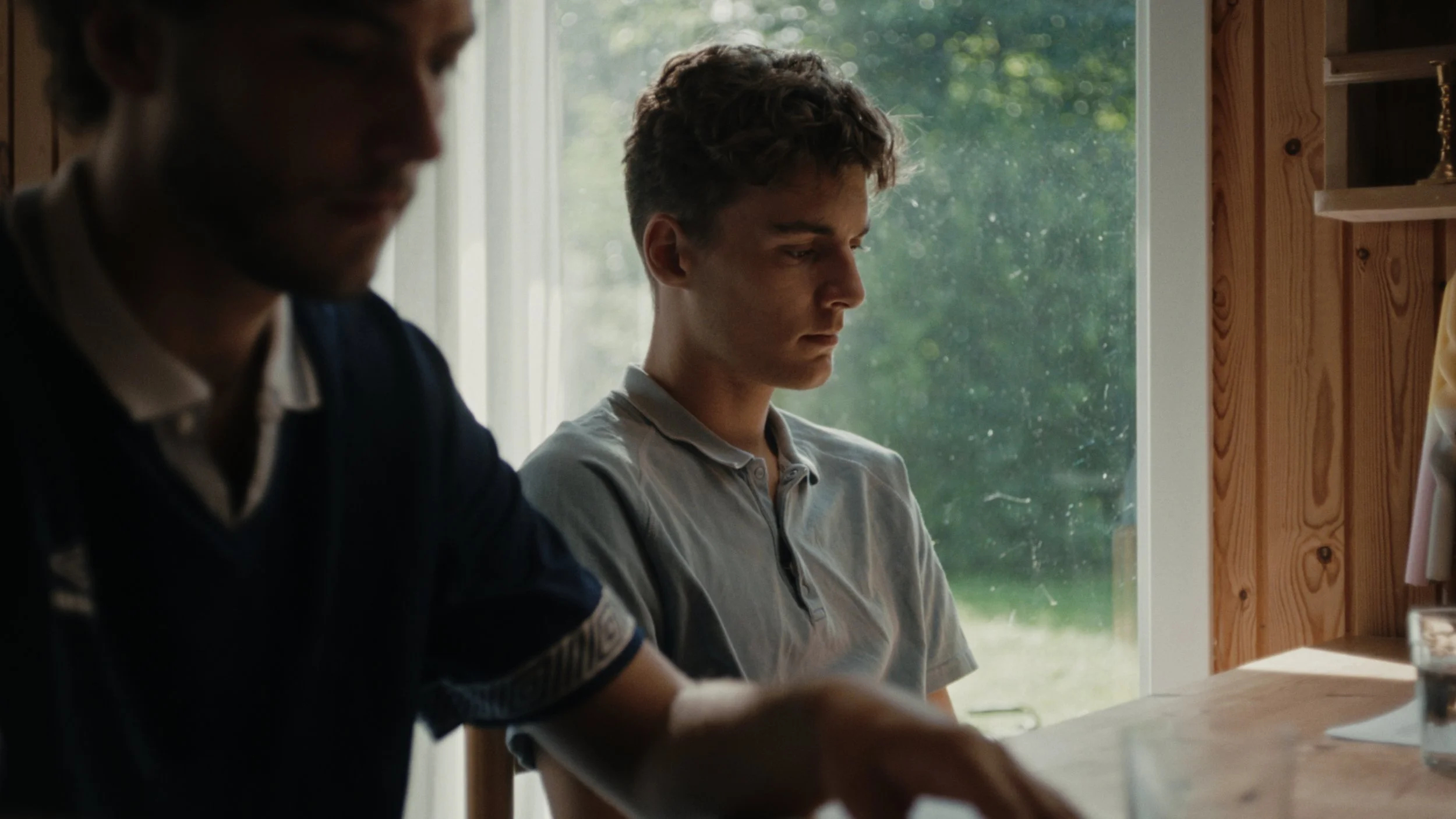 Two young men sitting indoors near a window. One is near the camera and blurred out, the other is in focus, looking down thoughtfully.