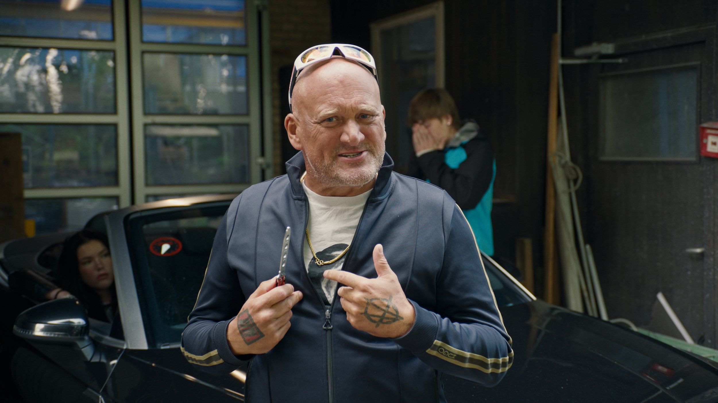 A man with tattoos, a gold chain, and a jacket holding a small knife, standing in a garage with a black sports car and two people in the background.