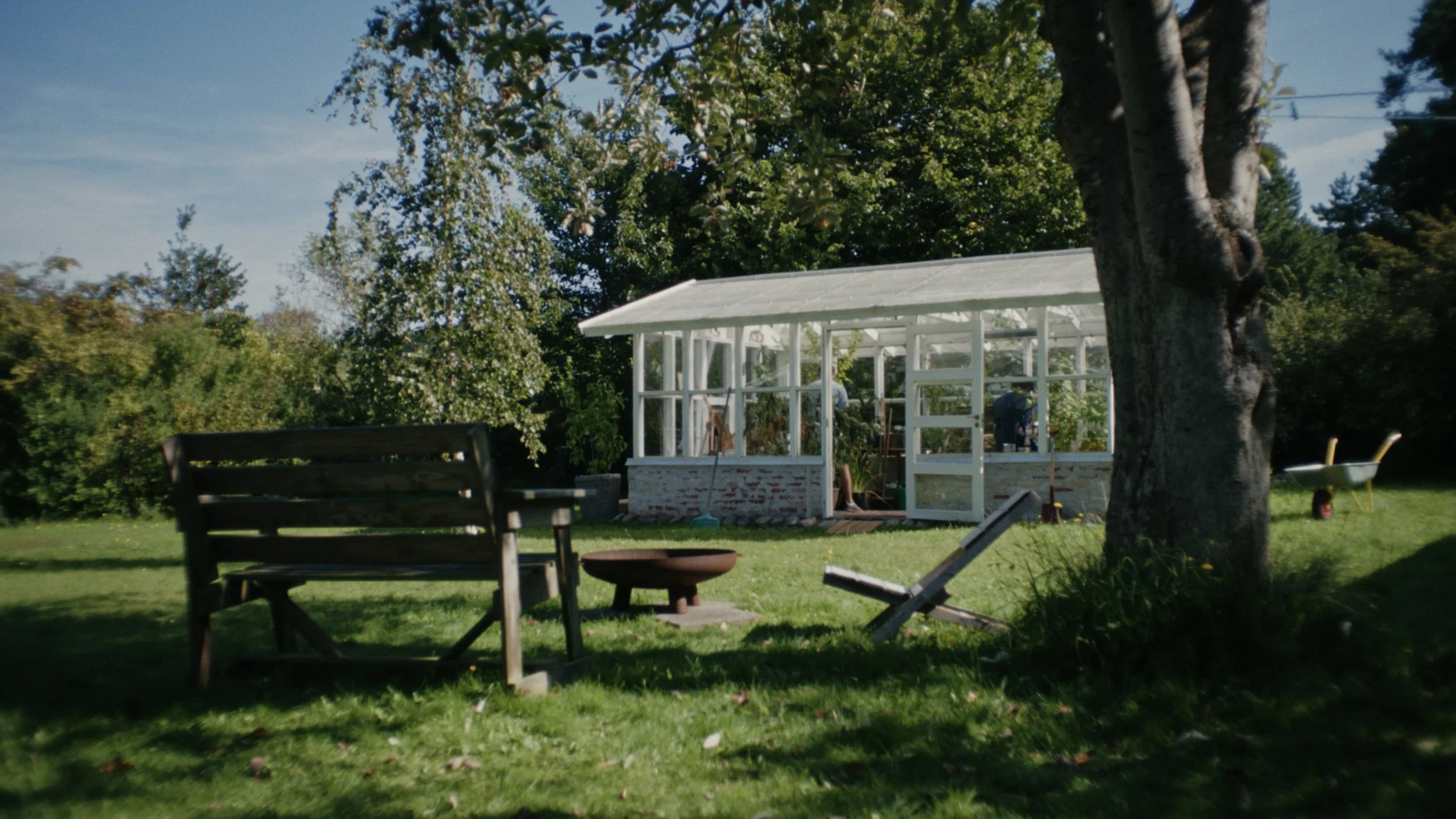 A backyard scene with a wooden bench, a fire pit, and a greenhouse under a tree, with gardening tools and a wheelbarrow nearby.