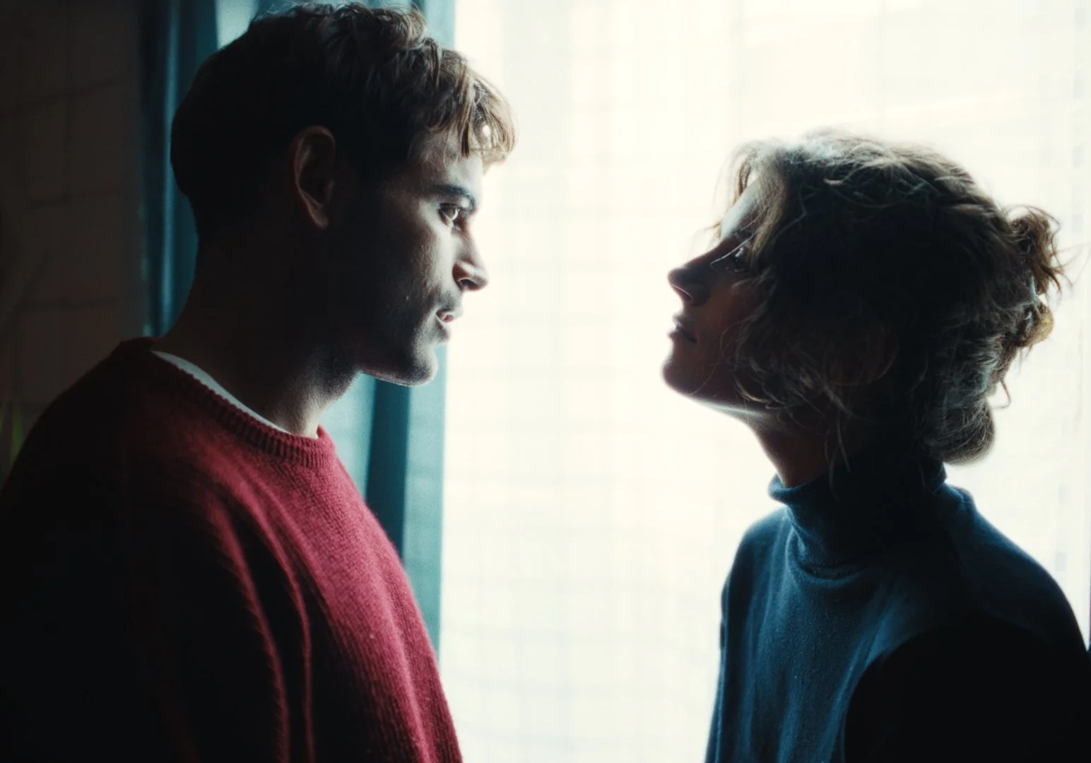 Two people, a man in a red sweater and a woman in a blue shirt, standing close and facing each other indoors near a window with sheer curtains, with their faces nearly touching.