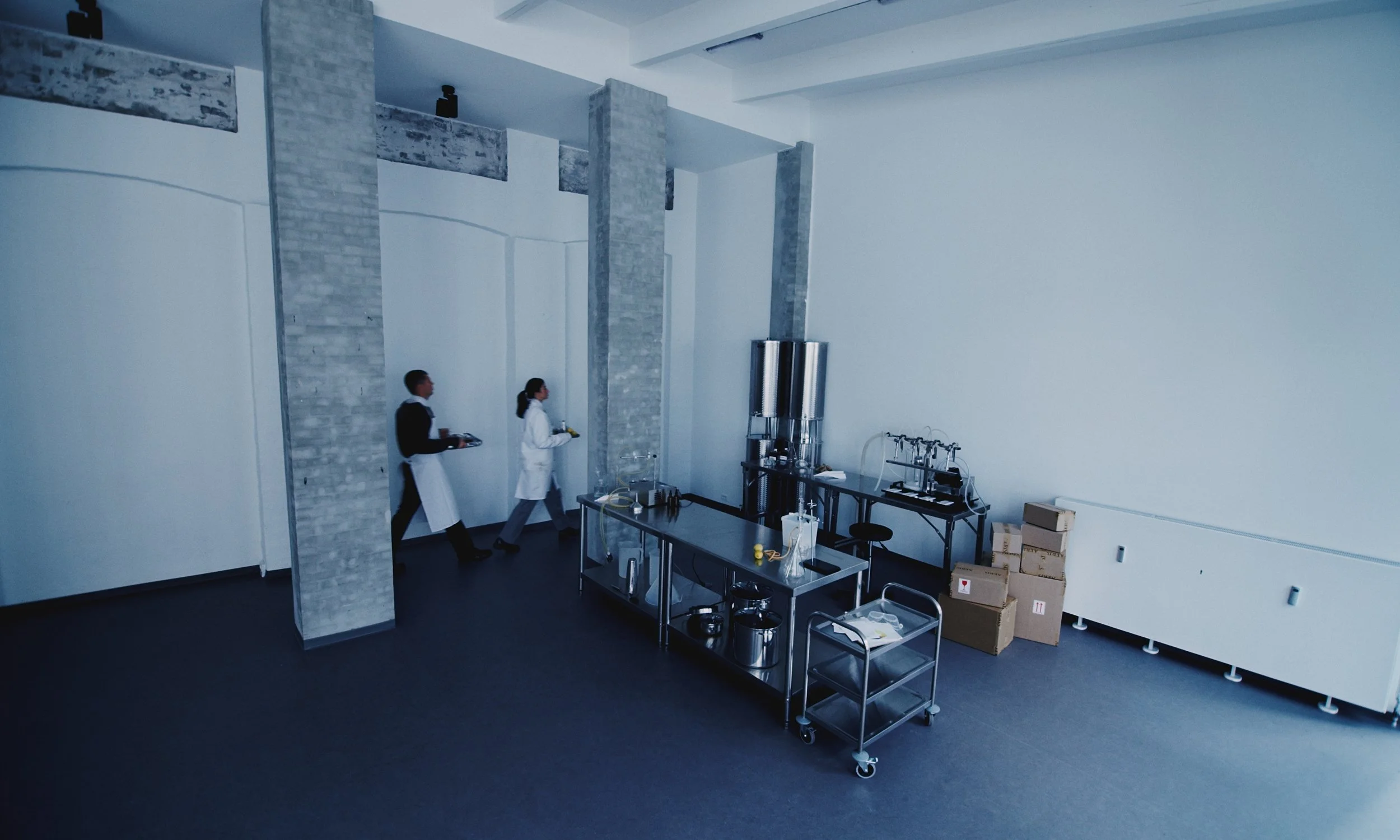 Laboratory or industrial workspace with three individuals walking in a line, a stainless steel table with lab equipment, stacked cardboard boxes, and large metal containers against a white wall with inclusion of brick columns.