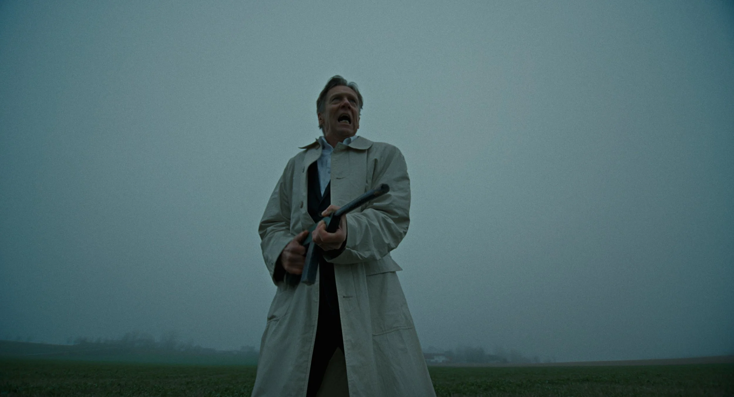 Man in beige trench coat holding a crowbar, standing in a grassy field with a cloudy, overcast sky, appearing distressed or yelling.