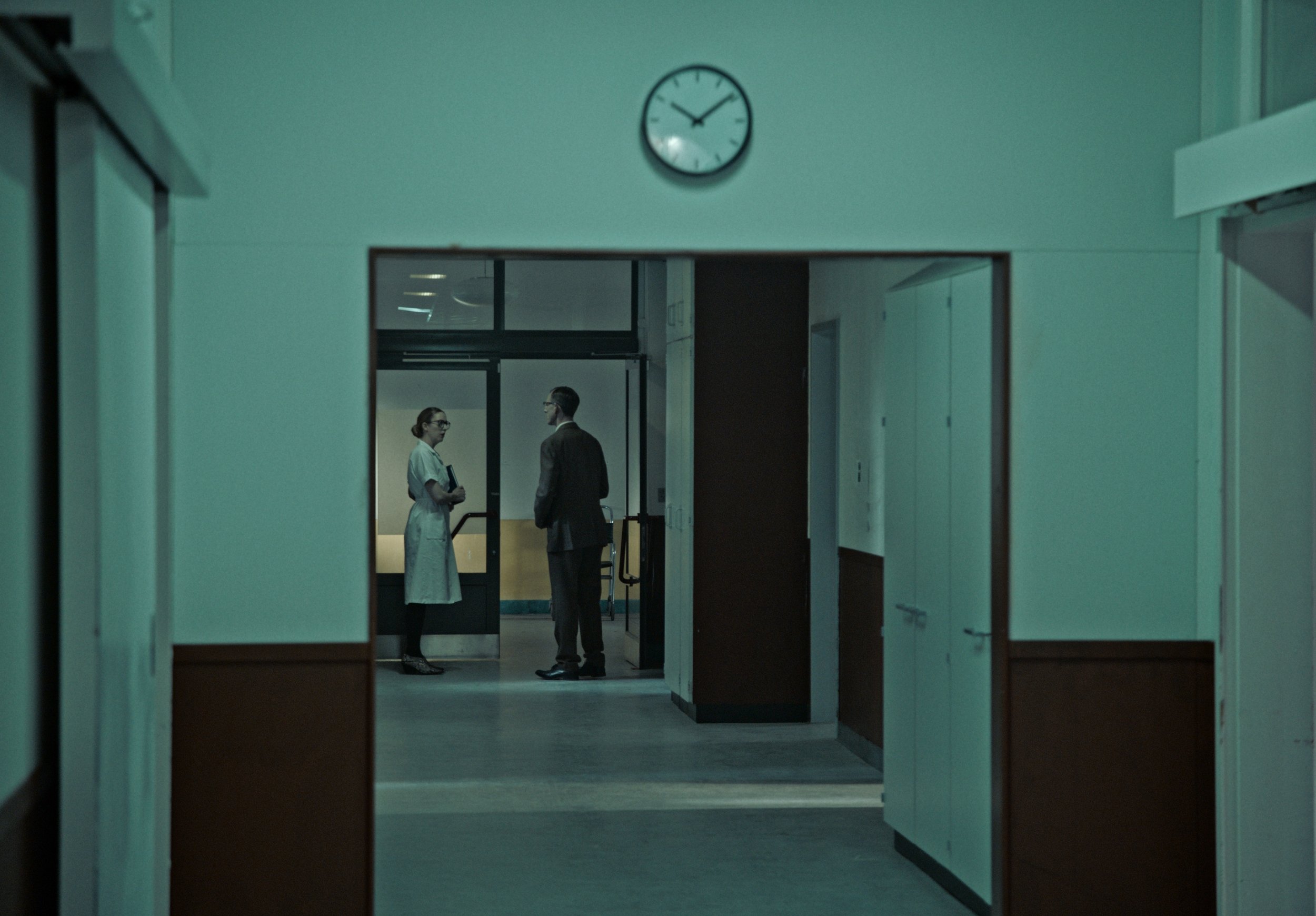 Hospital corridor viewed through an opening, with a clock showing 3:55, and two medical professionals engaged in conversation in the background.