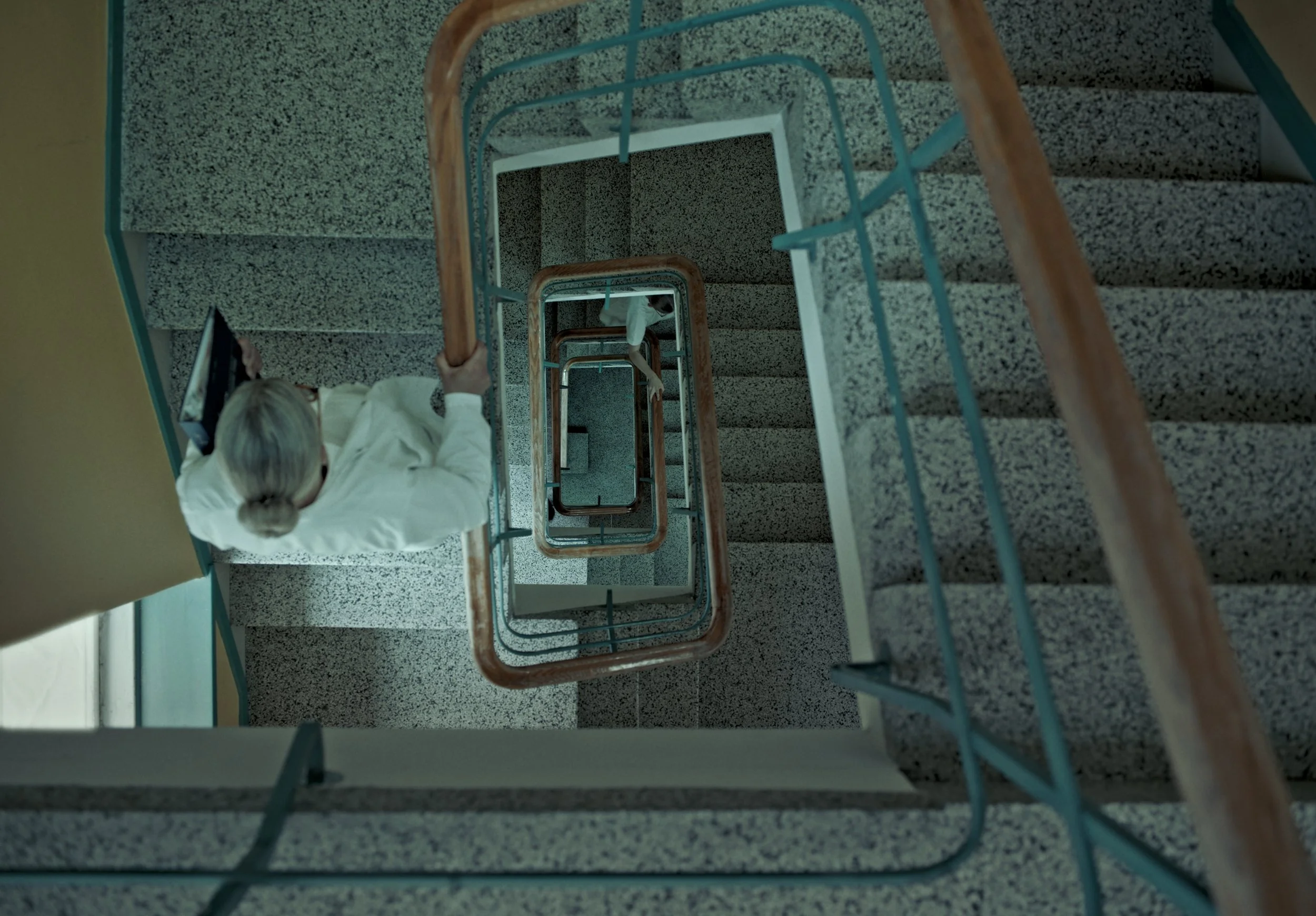 A top-down view of a stairwell with patterned gray steps and blue railings, taken from the upper floor looking downward.