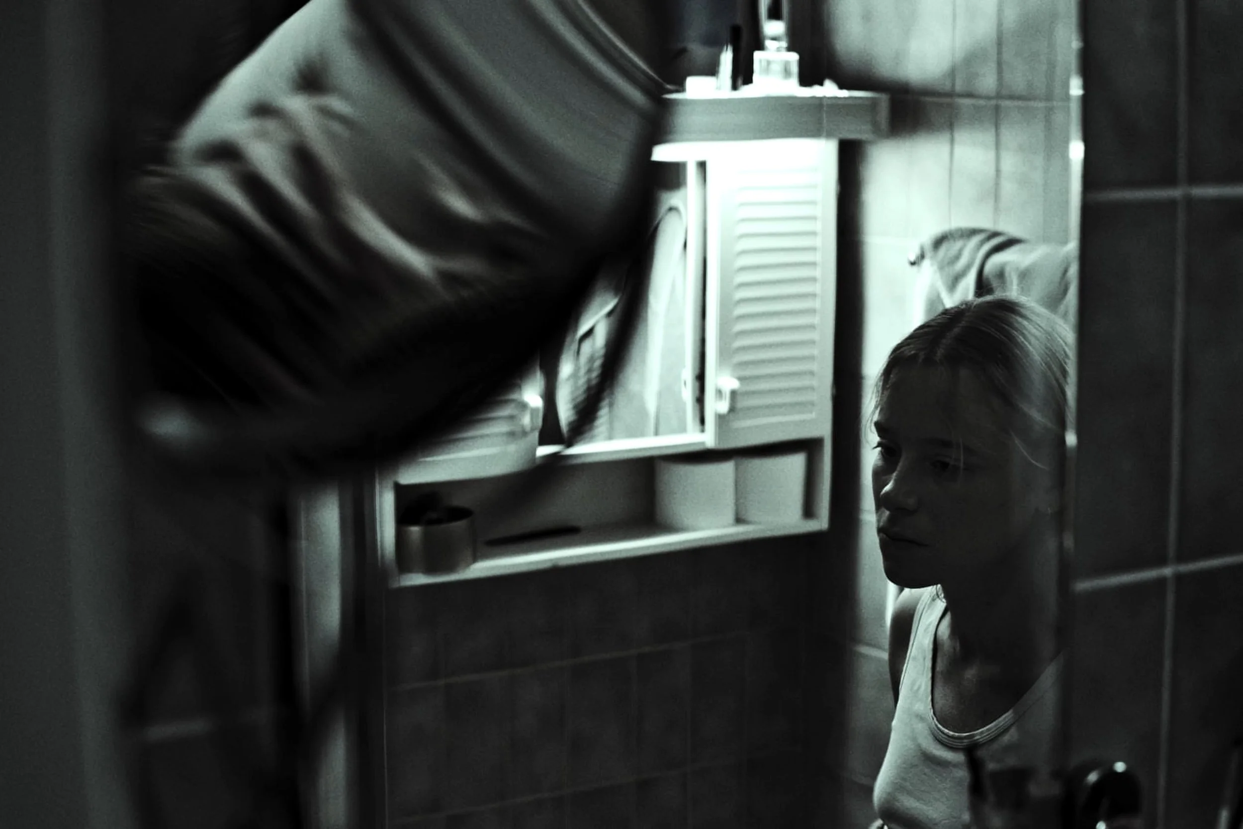 Black and white photo of a girl looking at her reflection in a mirror, with a bathroom cabinet partially visible.