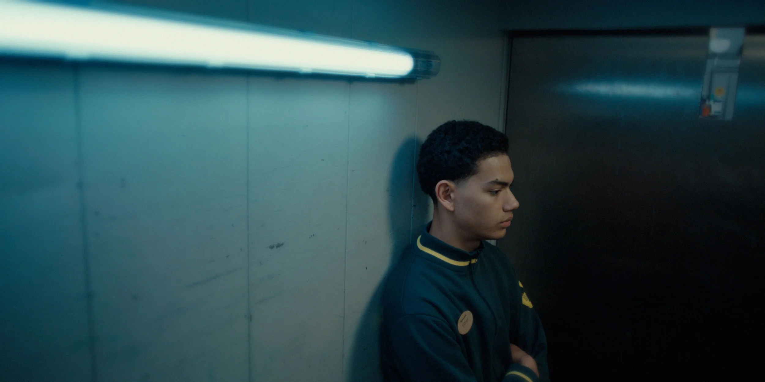 A young person with short curly hair, wearing a dark jacket with yellow accents, stands with arms crossed in a dimly lit room, leaning against a wall, with a fluorescent light above.