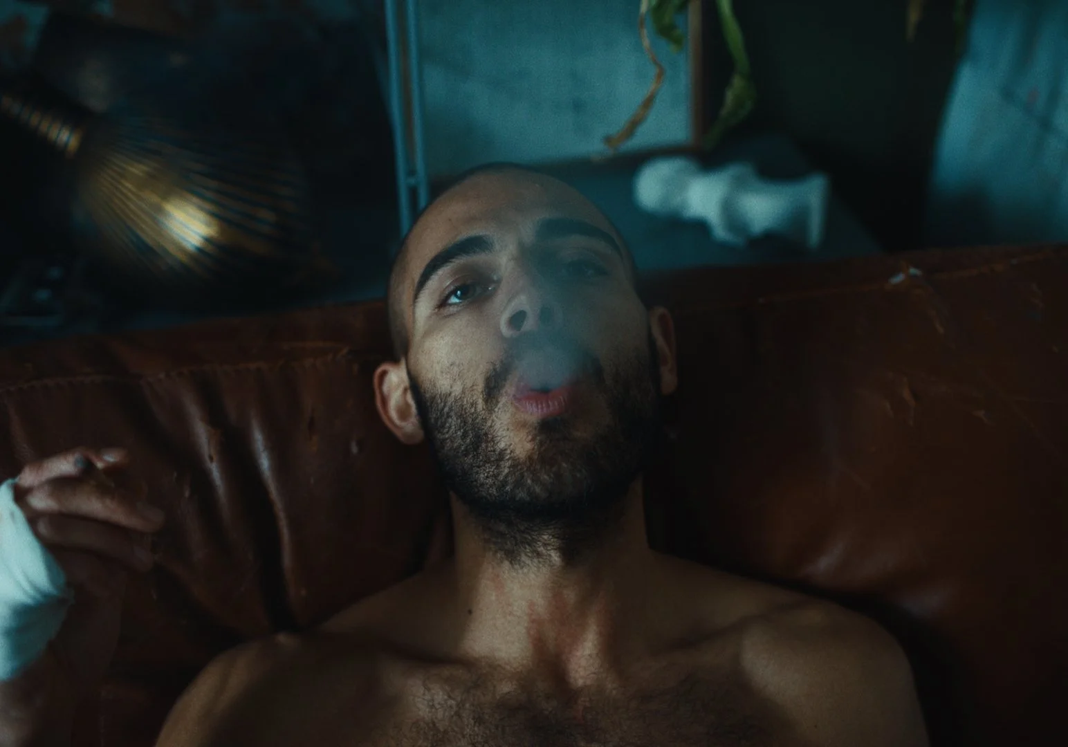 A shirtless man with a beard lies on a brown leather couch, smoking with smoke rising from his mouth, with a hand in a white glove holding a cigarette.