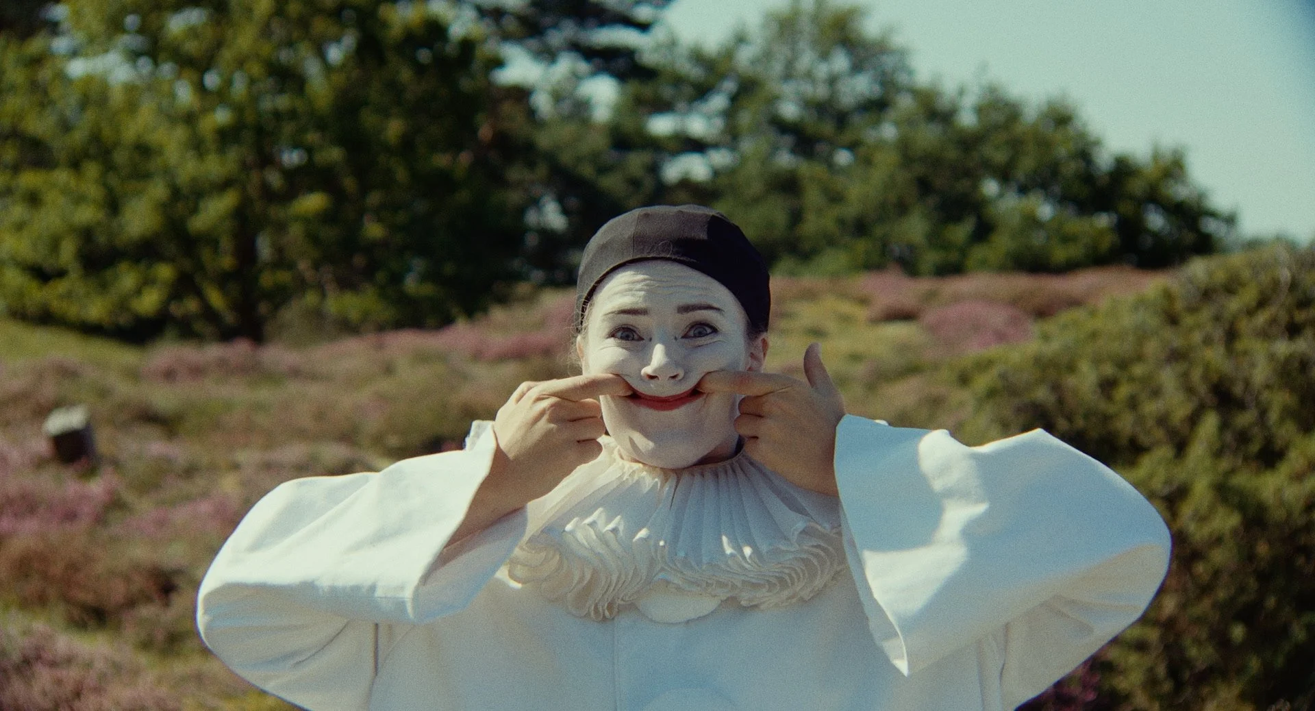Person dressed as a clown smiling and pulling their mouth into a smile with hands outdoors on a sunny day.