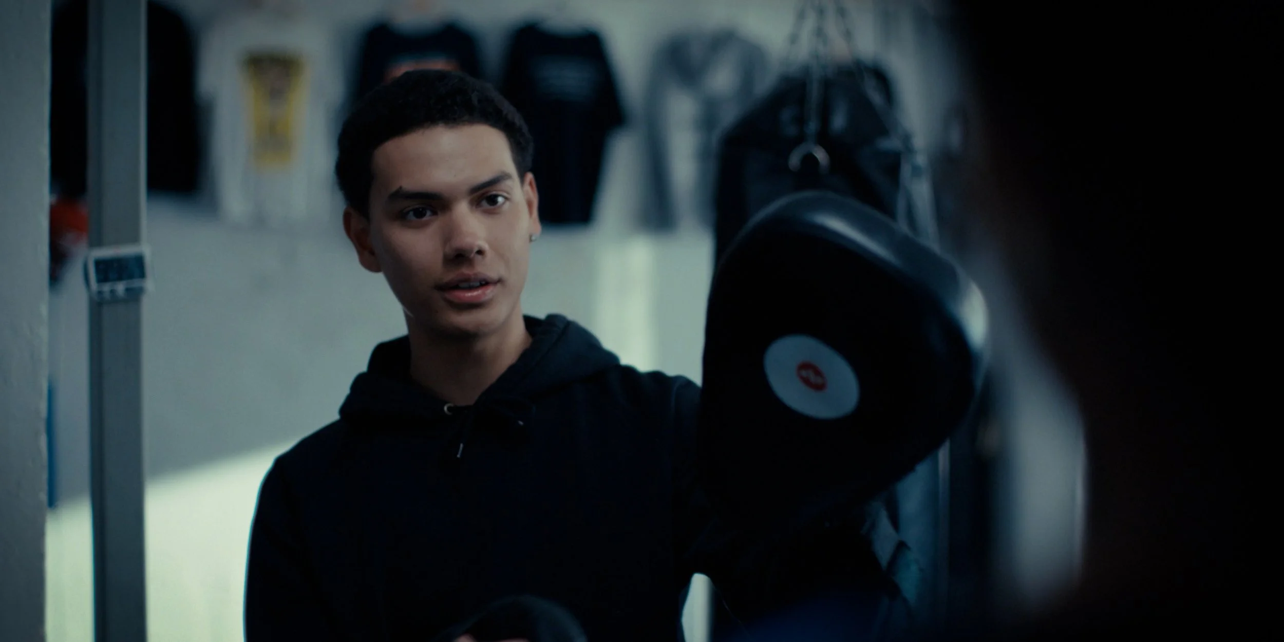 A young man in a black hoodie training with boxing gloves in a gym, with punching bags hanging in the background.
