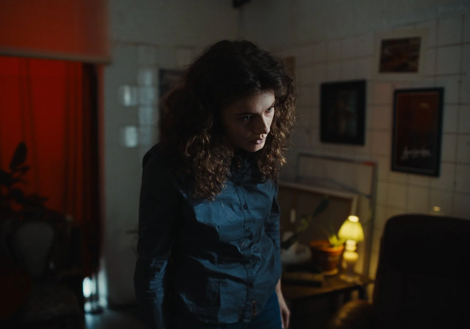 A woman with curly brown hair looking down in a dimly lit room with framed pictures on the wall and a small lamp on a table.