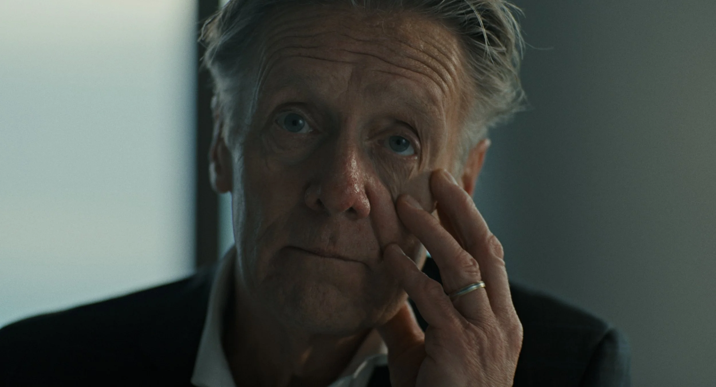 A close-up of an elderly man with gray hair and blue eyes, resting his face on his hand with fingers touching his cheek and eye, wearing a dark blazer and a white shirt, looking thoughtfully at the camera.