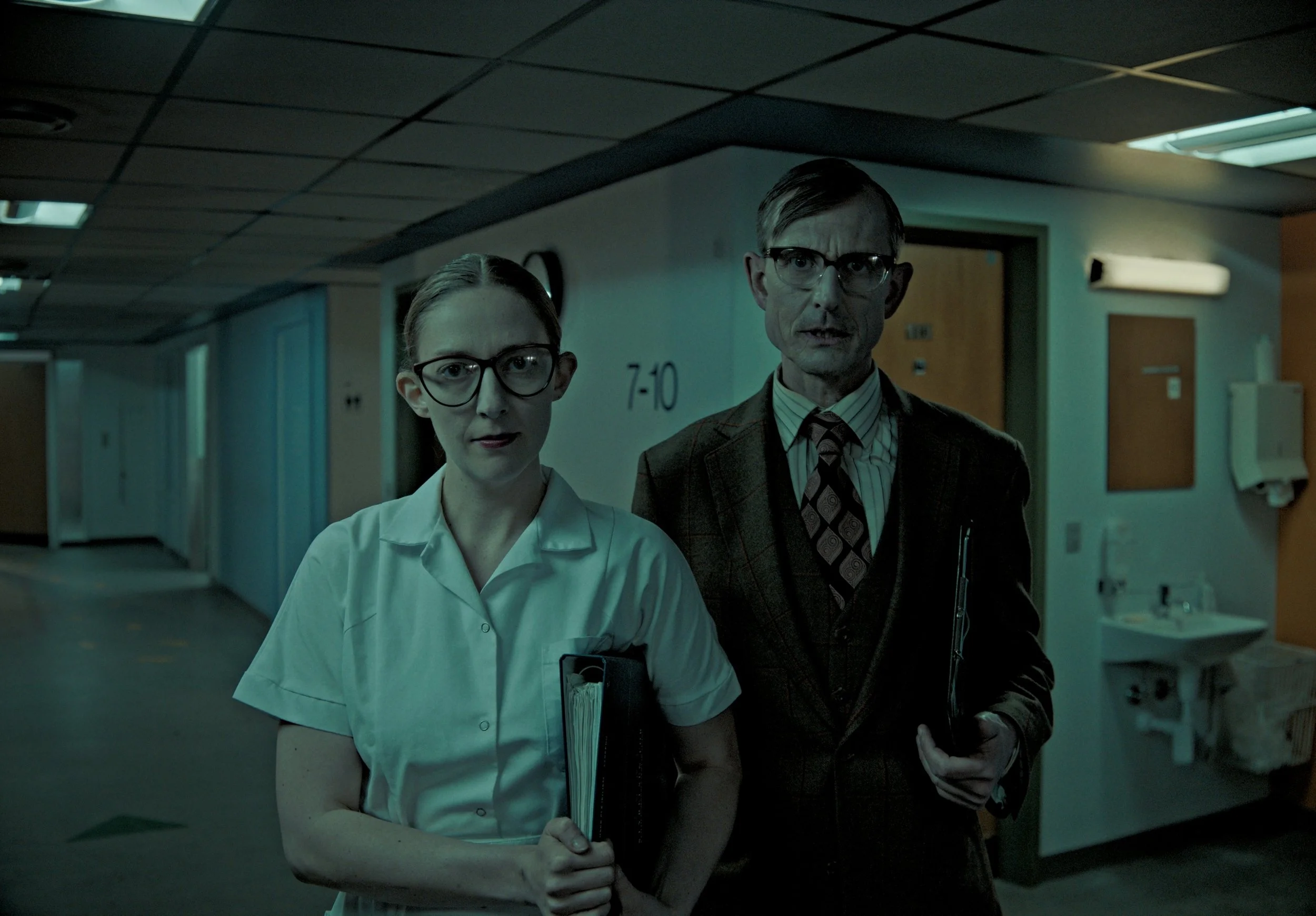 Two people, a woman dressed as a nurse and a man dressed as a doctor, stand in hallway with hospital or medical building background. Both wear glasses and hold folders or clipboards, facing forward with serious expressions.
