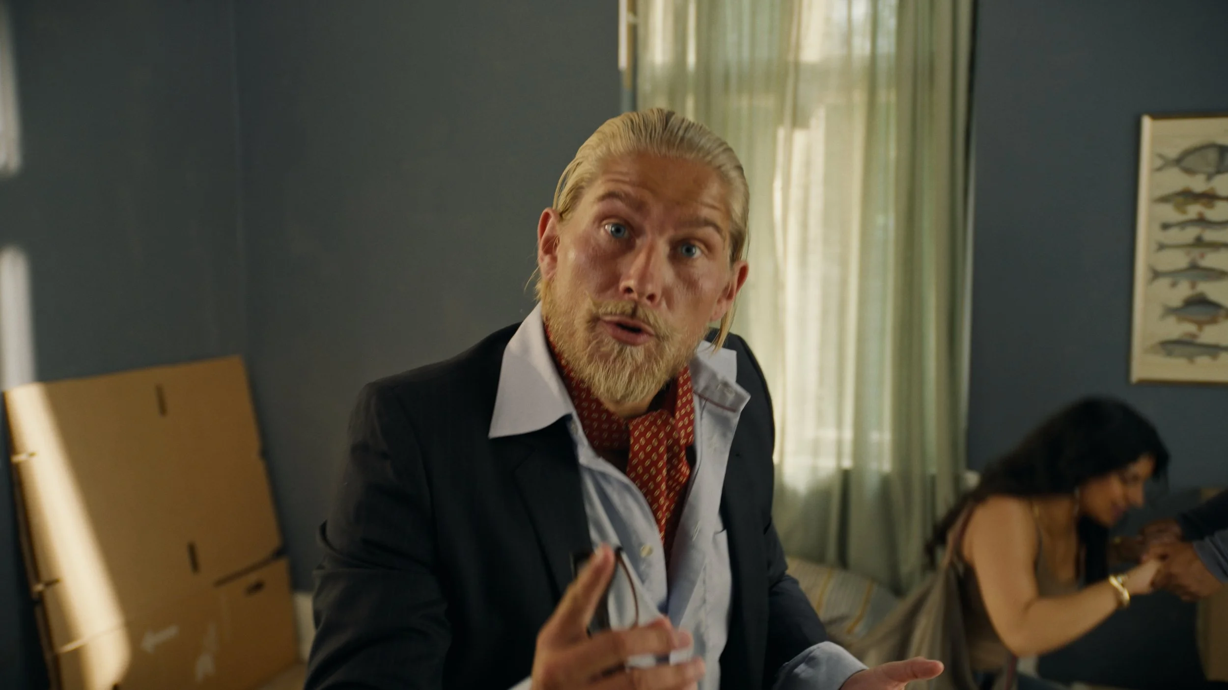A man with blonde hair and a beard, wearing a dark suit and a red patterned scarf, gestures with his finger while speaking. Behind him, a woman is sitting on a couch, holding hands with another person, with a fish-themed picture on the blue wall and 