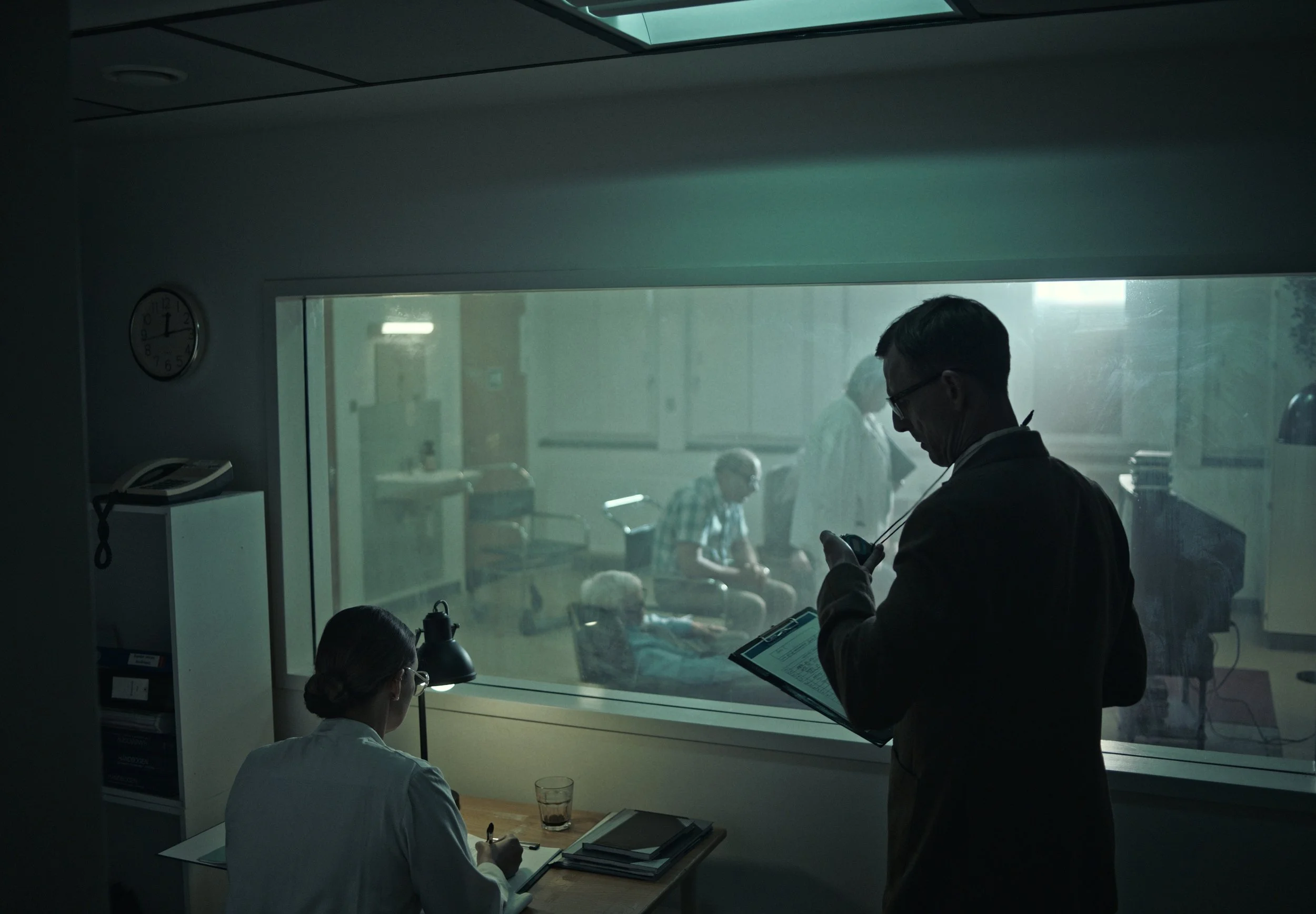 Scene inside a hospital or medical facility with two doctors or medical staff in foreground, one seated at a desk writing notes, the other standing and looking at a mobile device with a clipboard. Behind a large glass window, several patients are seated in a waiting area or lounge.