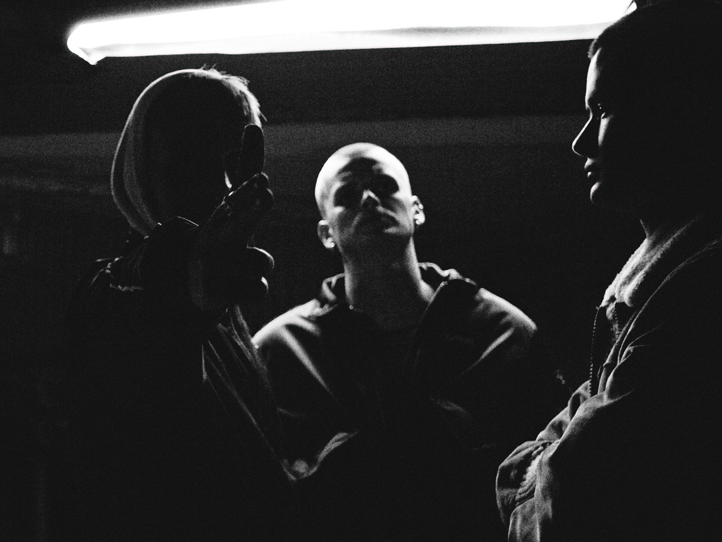 Three people in a dark setting with dramatic lighting, one person facing the camera with a bald head, and the other two with faces in profile.