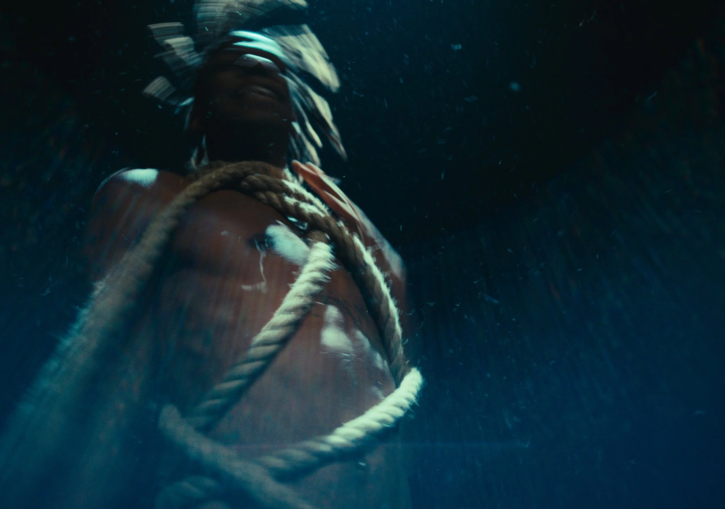 A person with braided hair underwater, wearing a brown jacket and a white rope around their shoulder, with water and bubbles surrounding them.