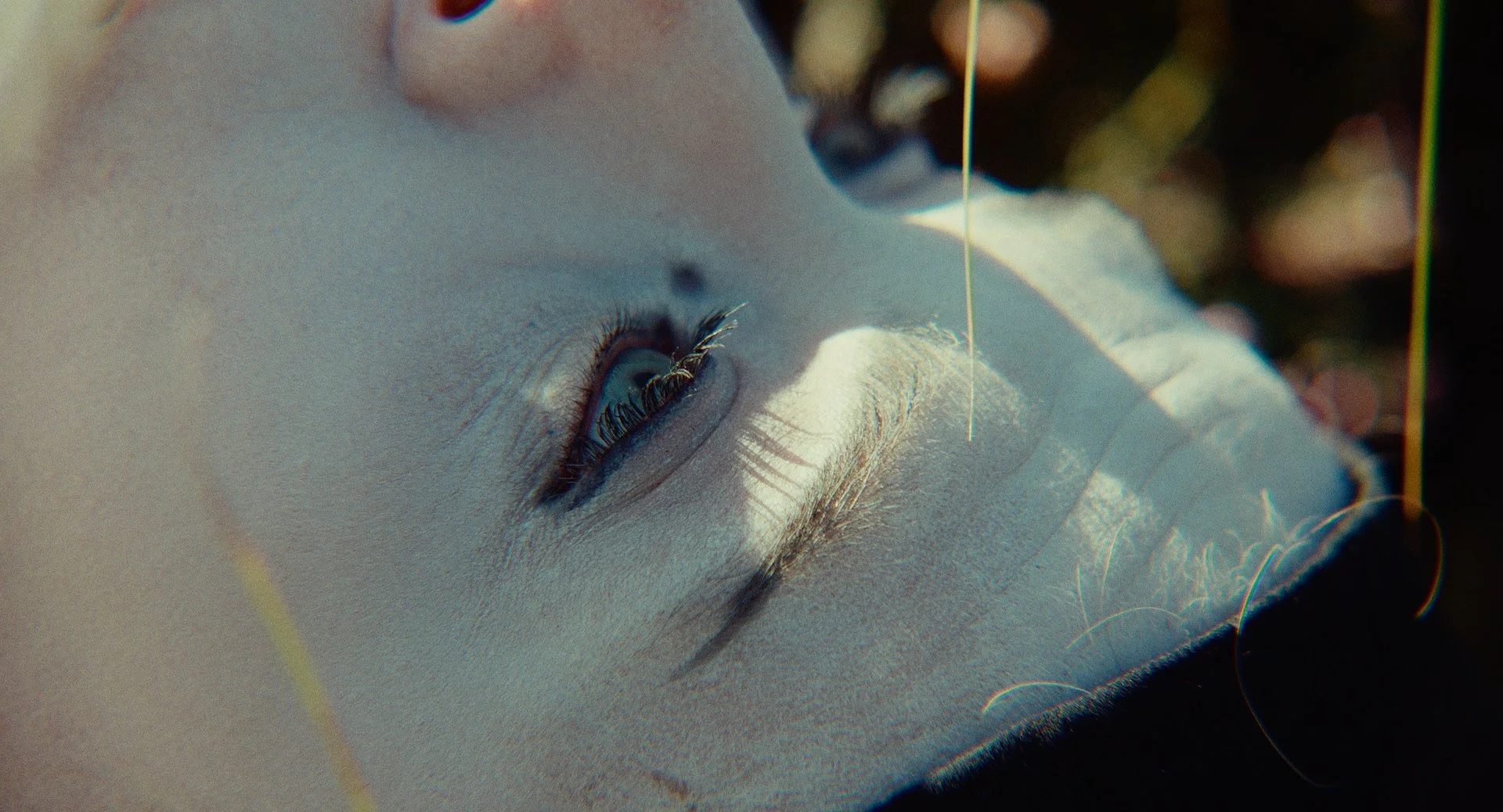 Close-up of a woman's face lying down outdoors, with sunlight casting shadows on her skin and her eyes partially open.