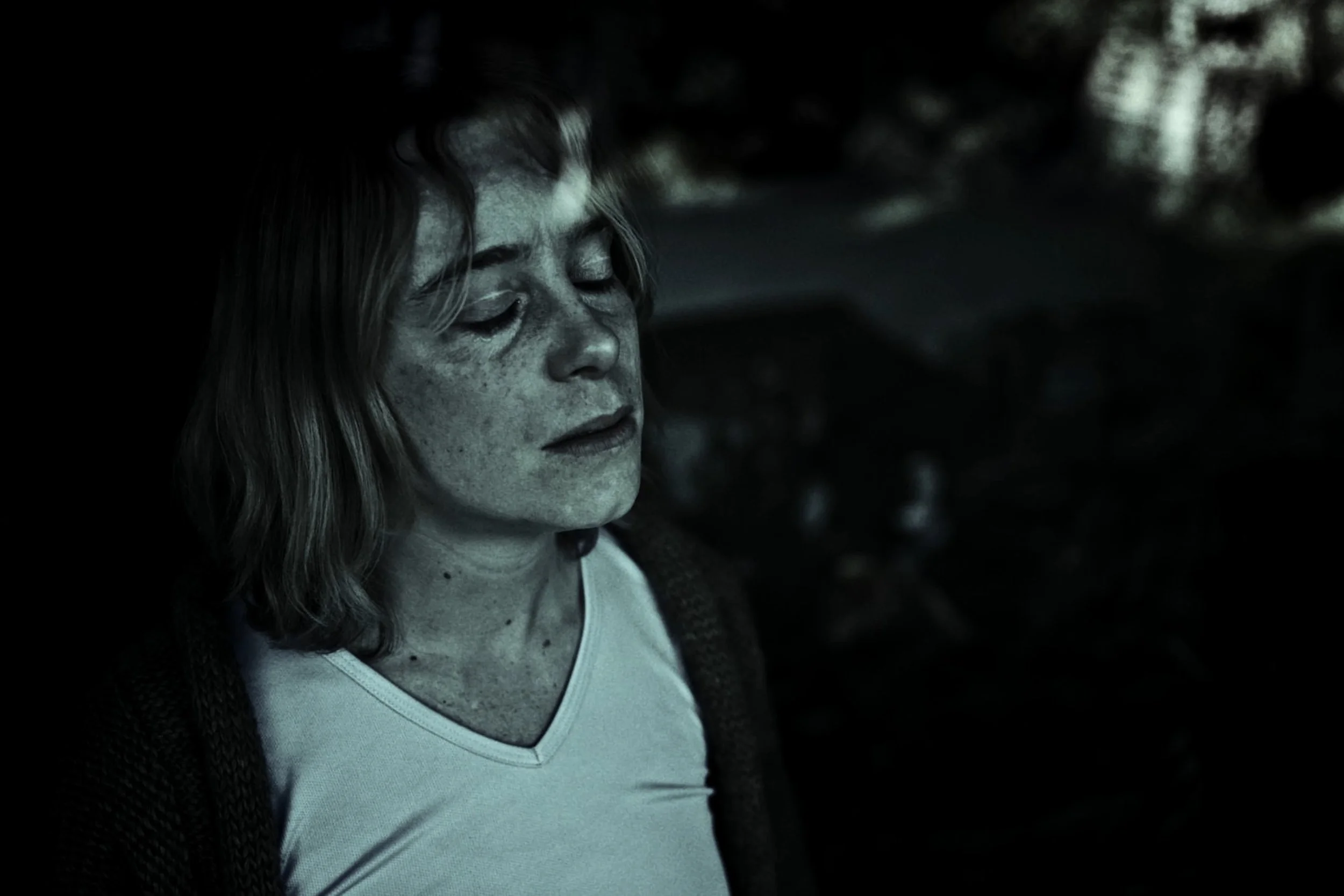 Close-up of a woman with short hair and freckles, eyes closed, appearing thoughtful or peaceful, in a dark environment.