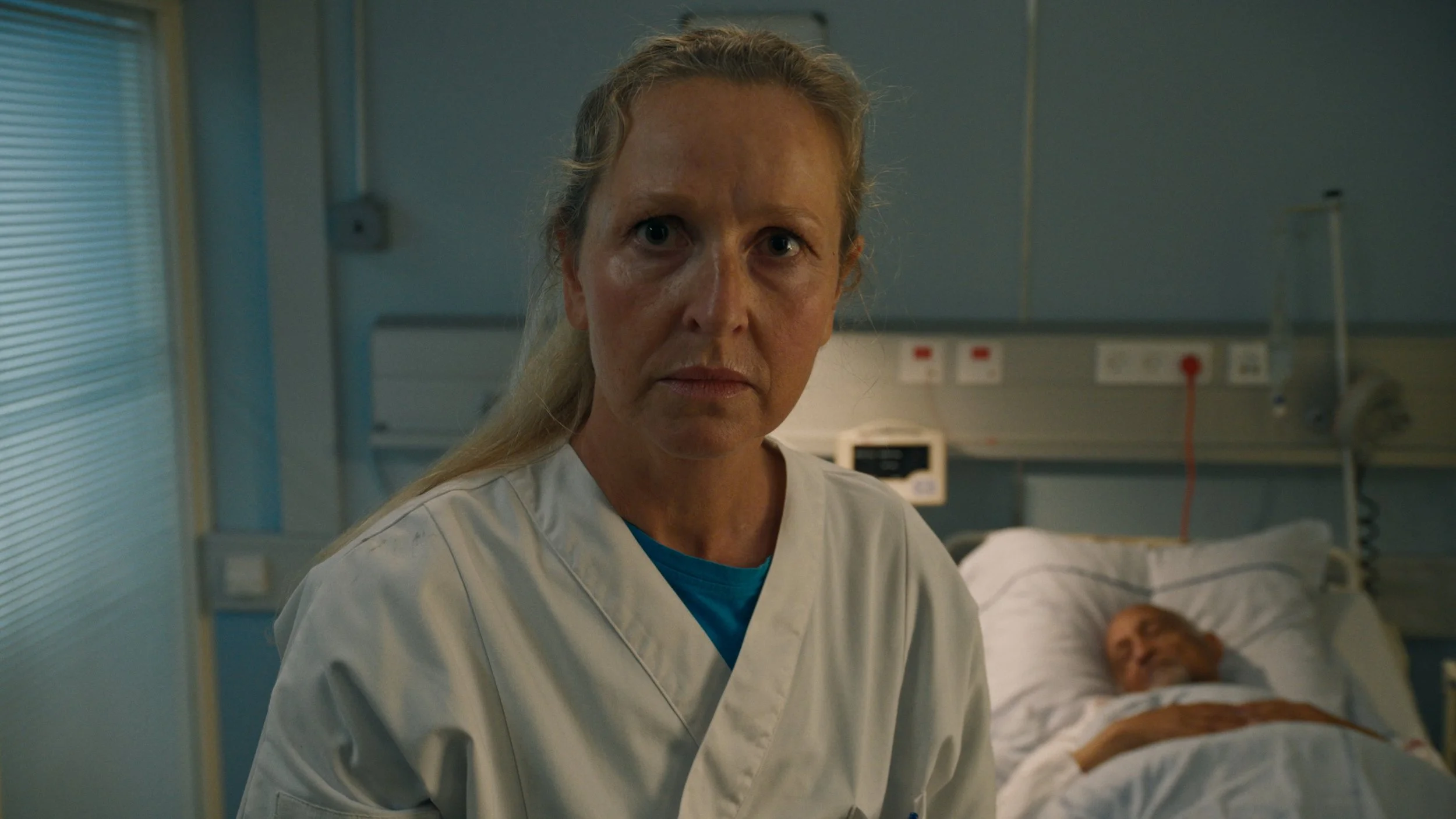 A serious-looking female nurse or doctor in a hospital room, standing beside a patient lying in bed. The patient appears unconscious or sleeping.