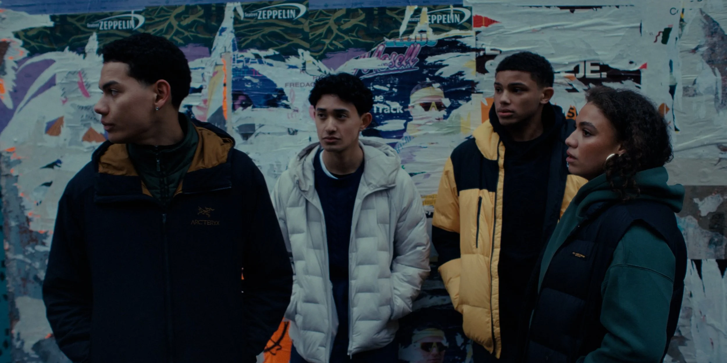 Four young people standing in front of a colorful, torn poster wall, wearing jackets, with one person looking to the side and the others looking forward.