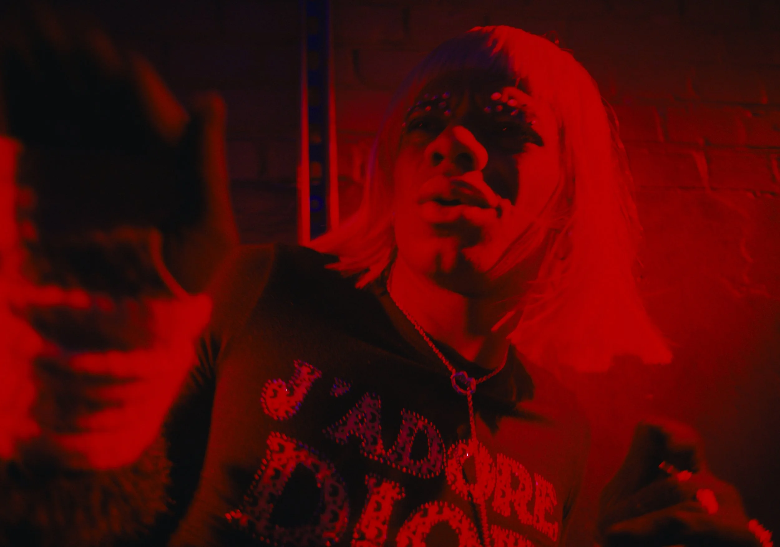 A person with bleached blonde hair and face paint taking a selfie in a dimly lit, red-tinted room, wearing a black shirt with the words 'Cadddrag' visible.