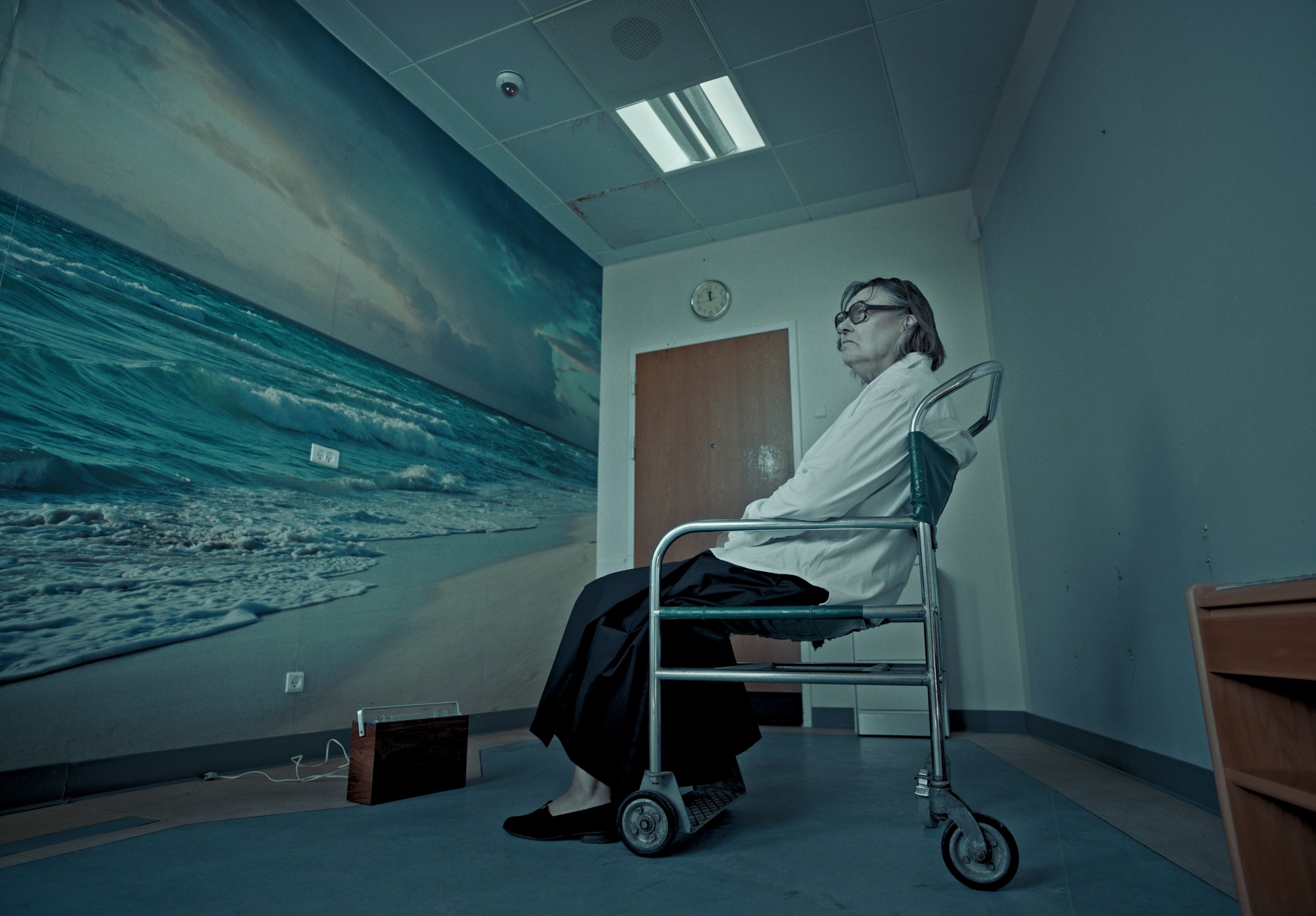 A woman sitting in a wheelchair, looking at a large mural of the ocean on the wall of a room, with a clock above the door.