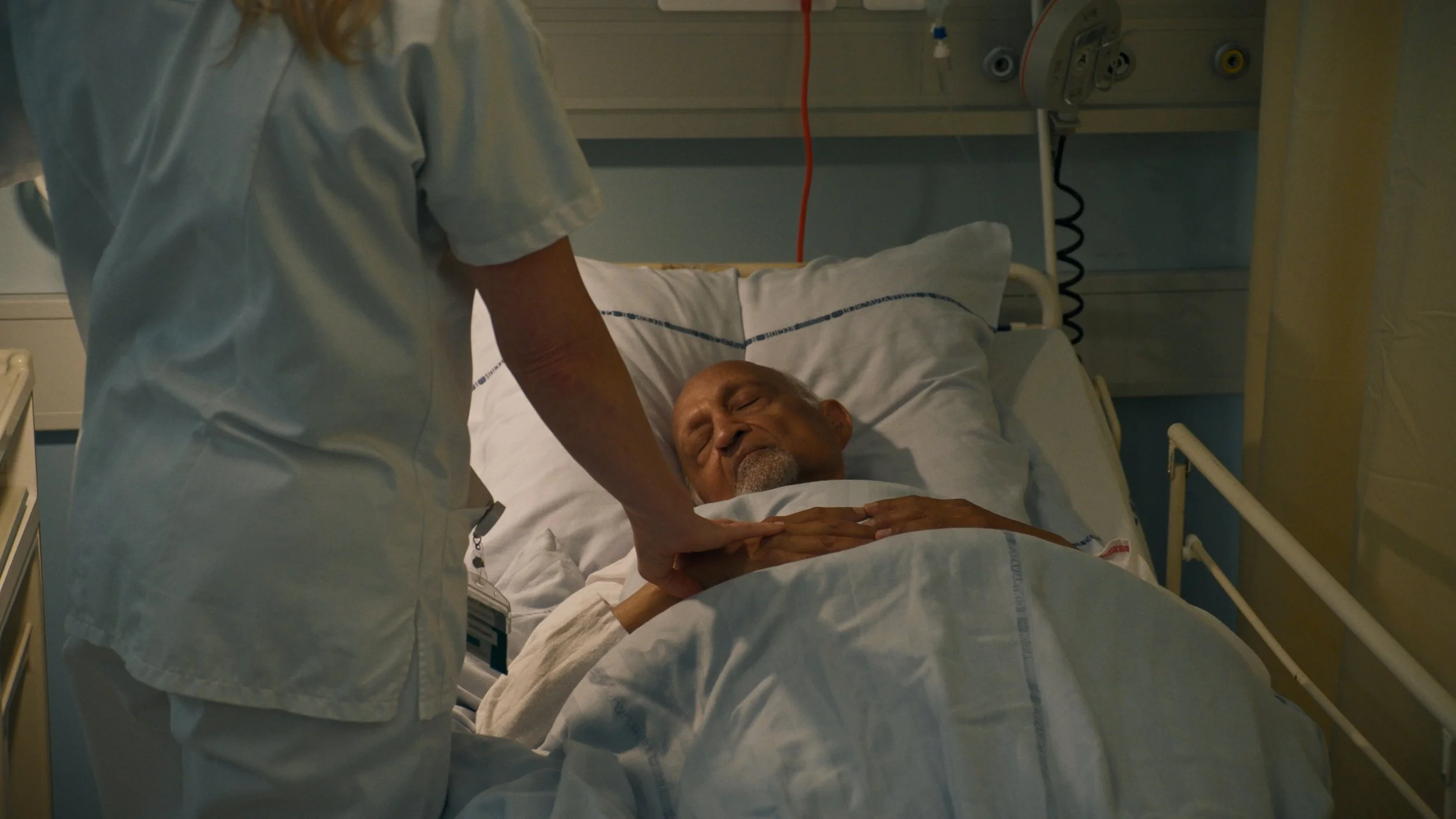 A nurse is checking on an elderly male patient lying in a hospital bed, holding his hand, in a hospital room.