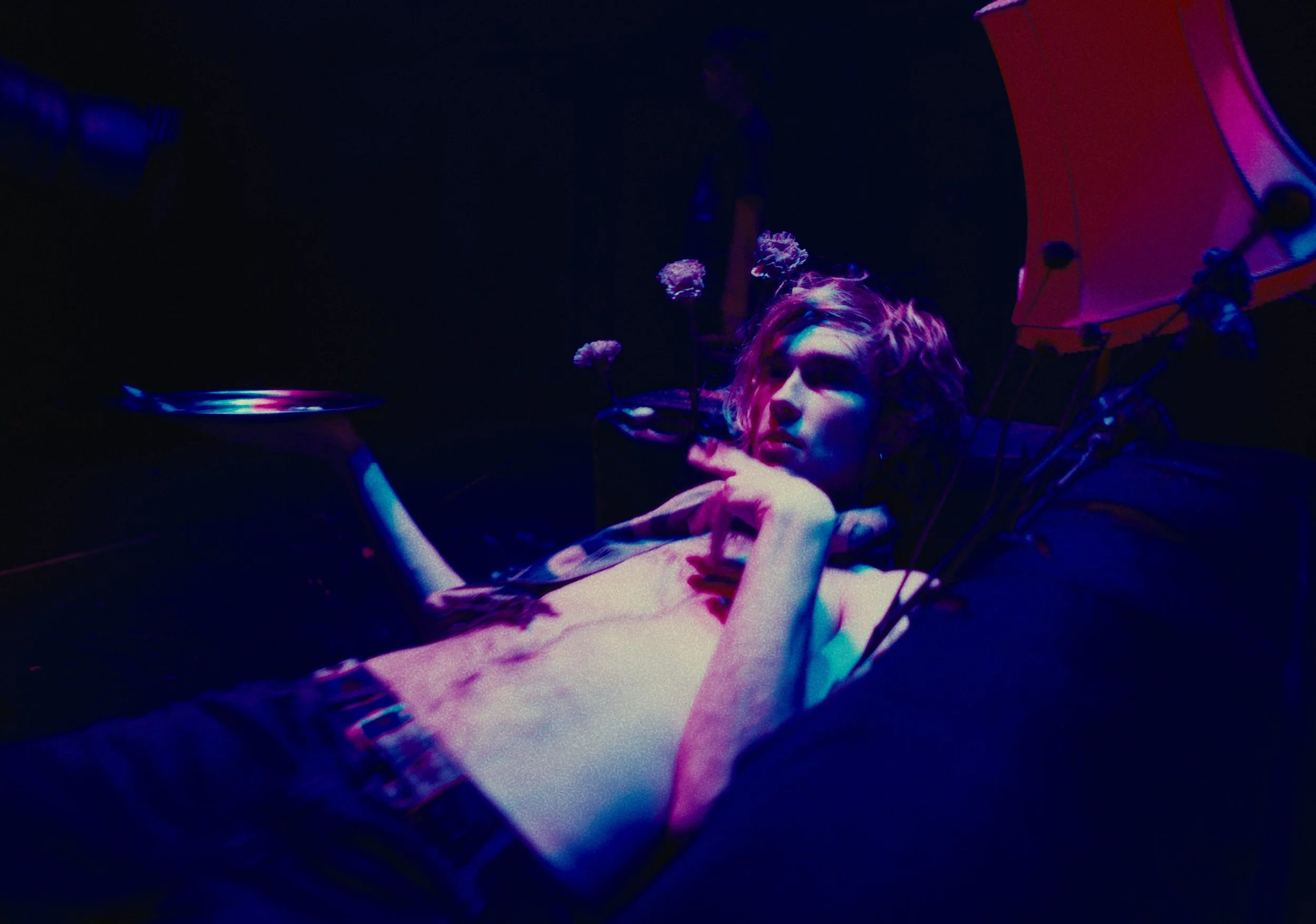 A woman with curly hair lying on a dark surface, illuminated by purple and blue lighting, holding a silver tray, surrounded by flowers and a pink umbrella