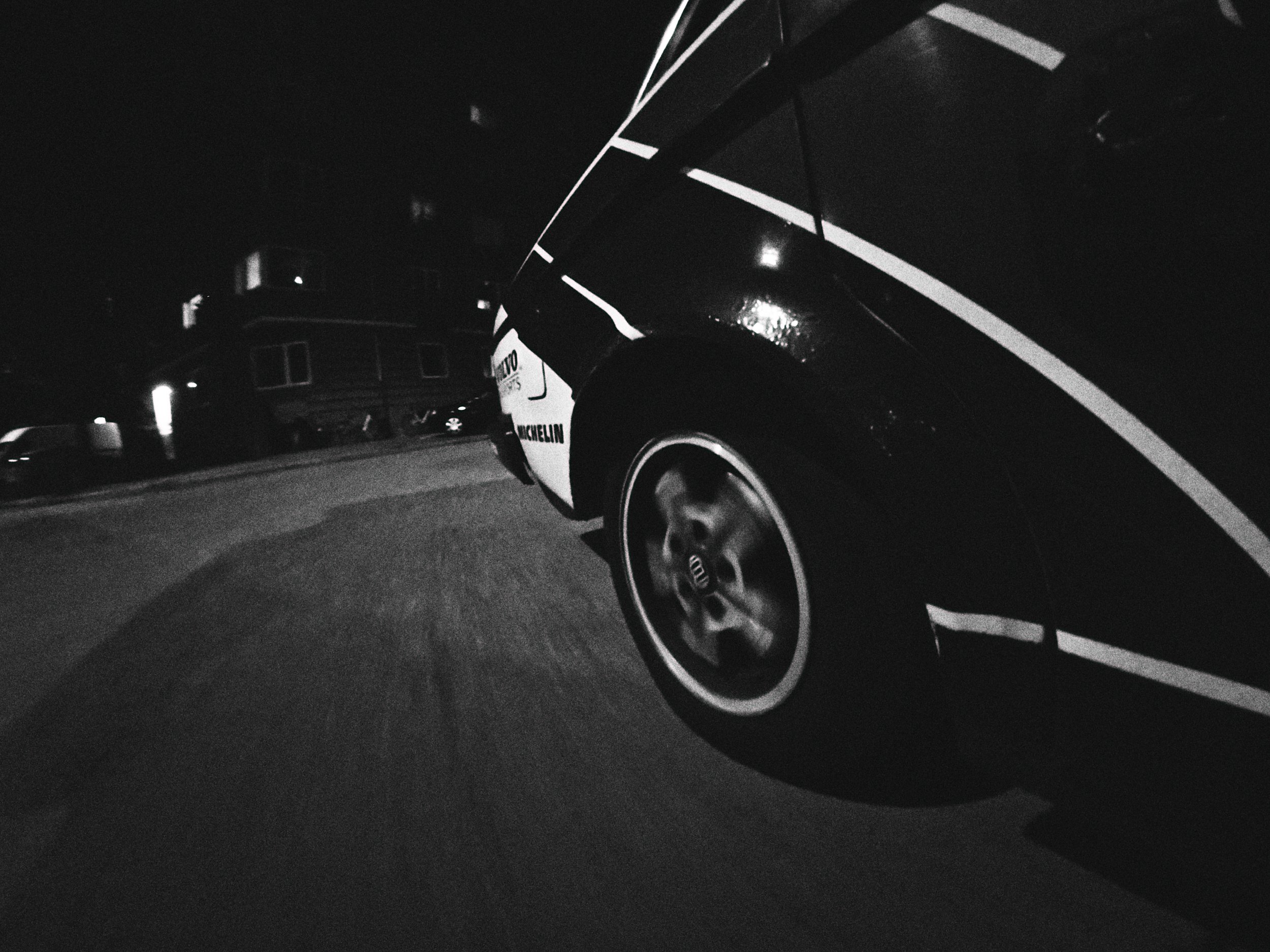 A black and white photo of a vintage race car with Michelin and Audi logos, taken at night, showing the front tire and part of the body, with a building and parked cars in the background.