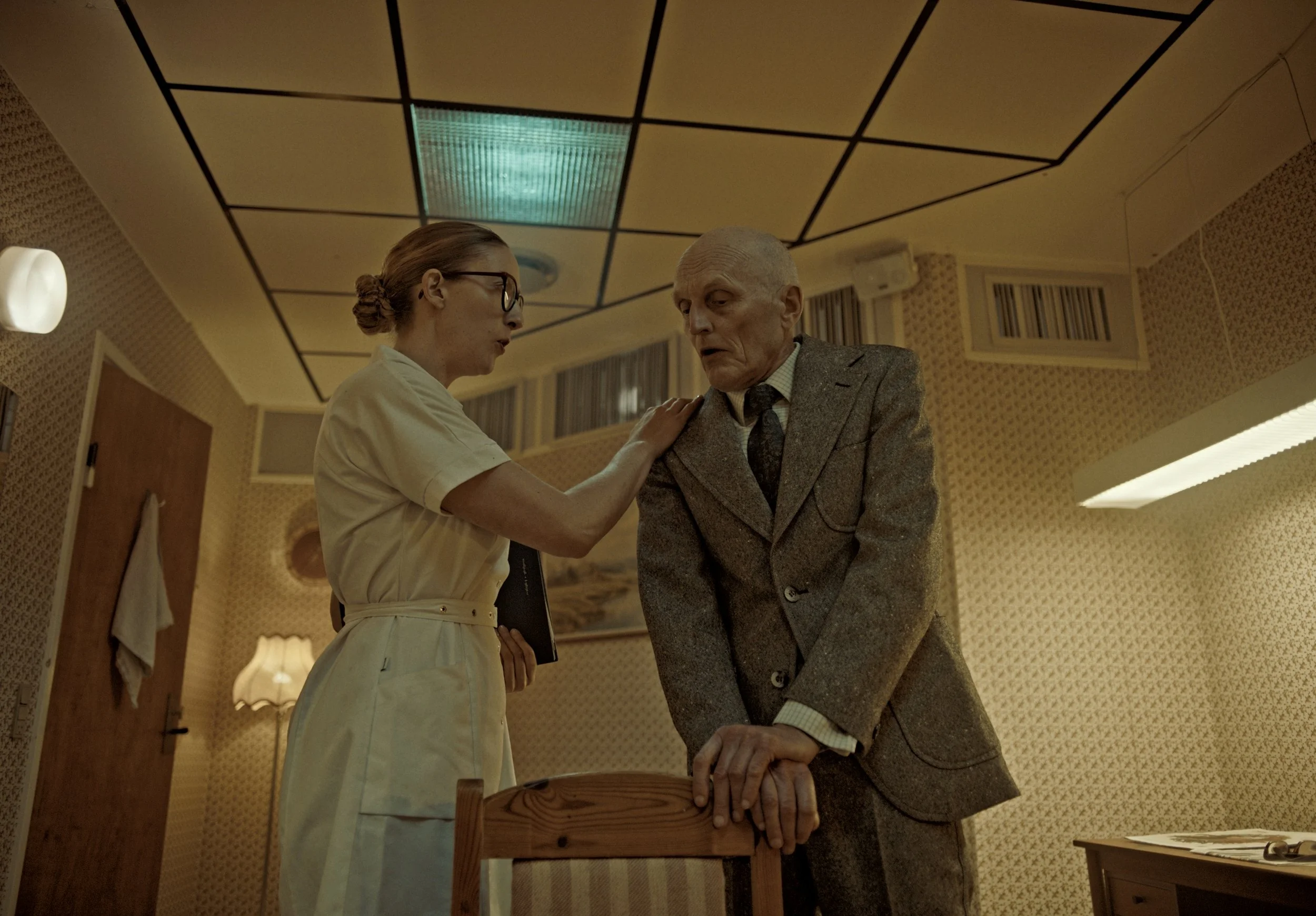 A woman in a nurse uniform comforting an elderly man in a gray suit in a patterned room, with a wooden chair and a lamp in the background.