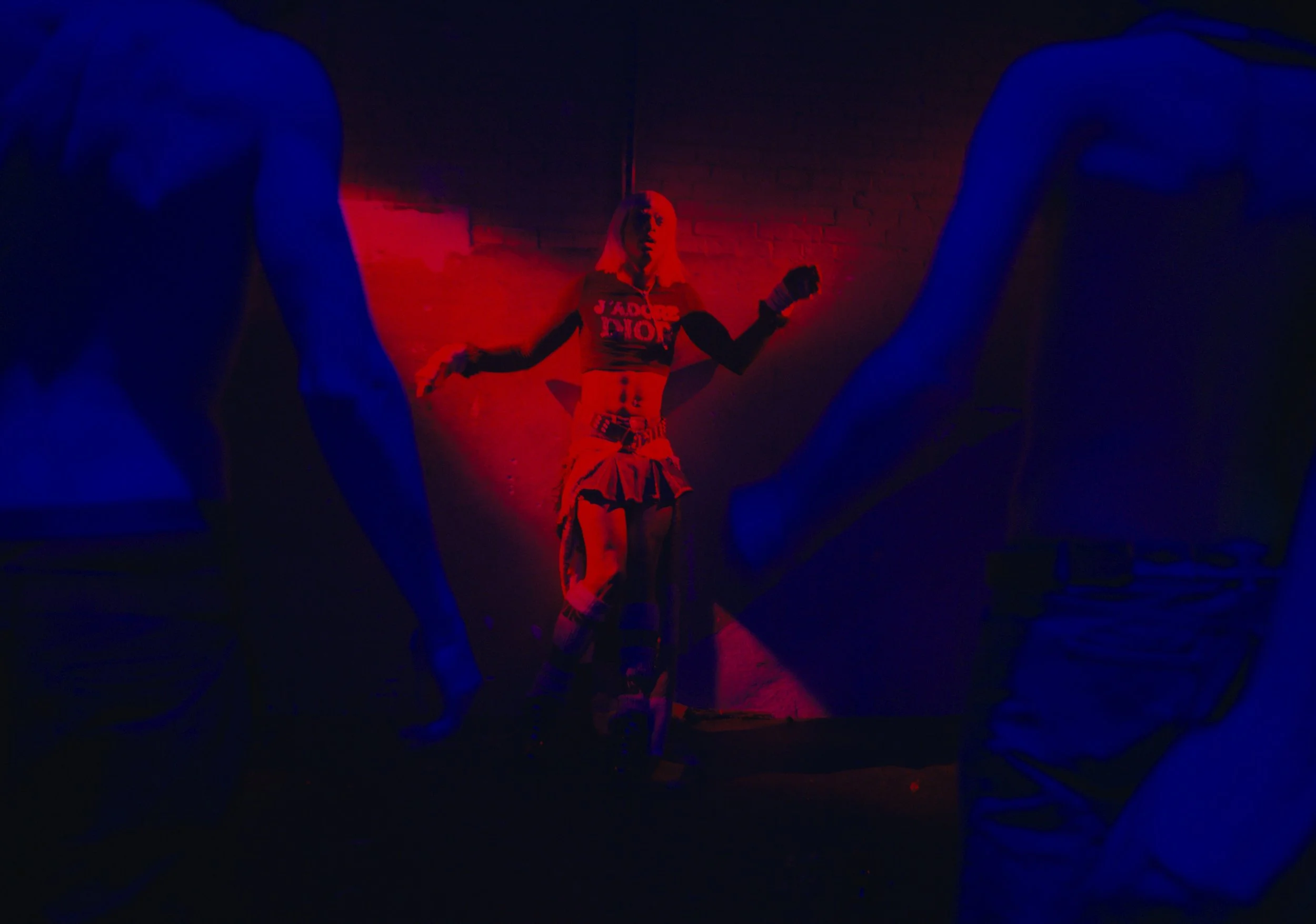 A woman with pink hair illuminated by red light, wearing a Dior crop top and a skirt, posed against a brick wall. Two muscular men are in the foreground, partially visible and in shadow, wearing sleeveless shirts.