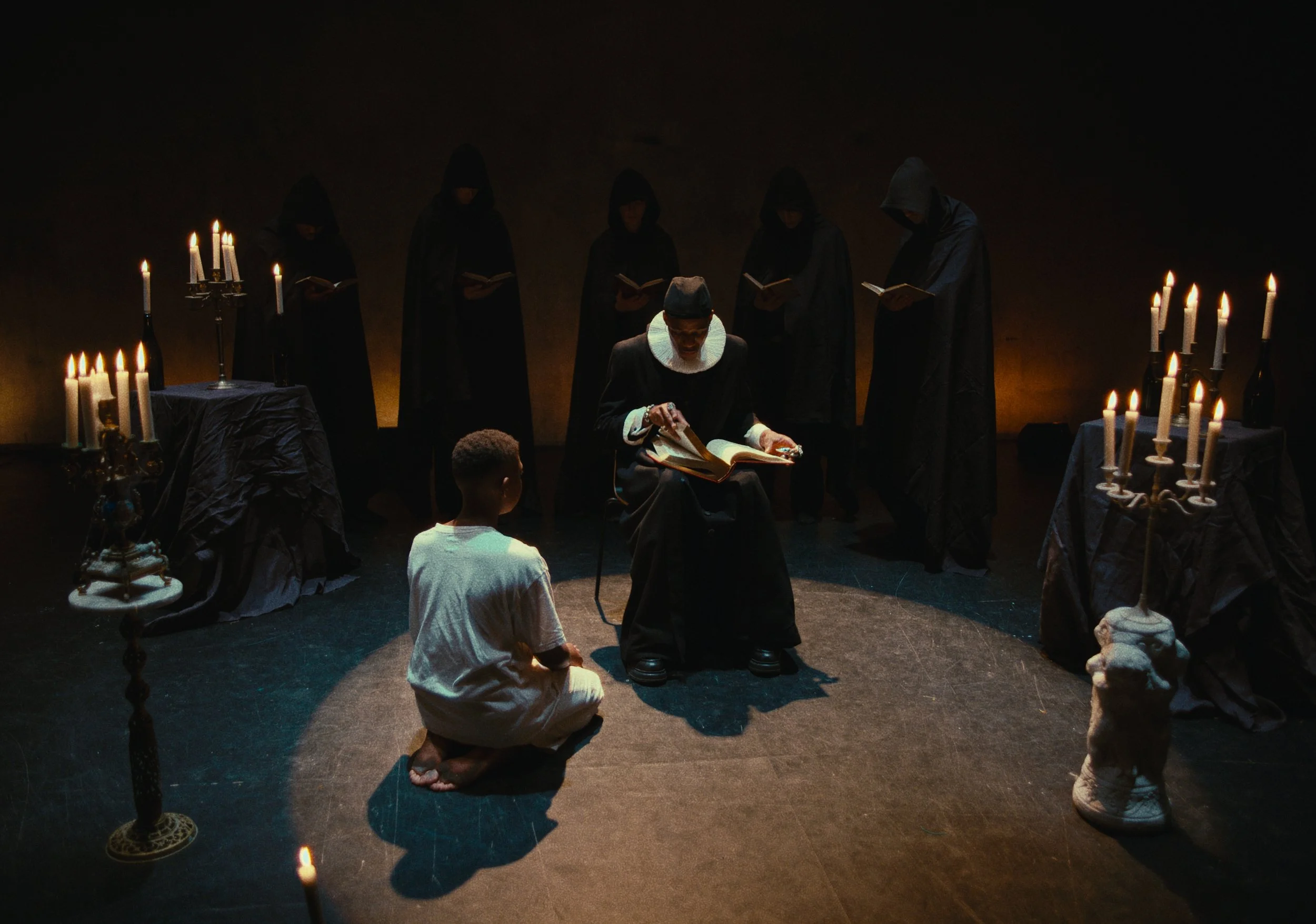 A person dressed as a nun sitting on a chair reading a book while a child in white kneels on the floor in front, surrounded by five figures in black robes with hoods, candles on ornate tables, in a dimly lit room.