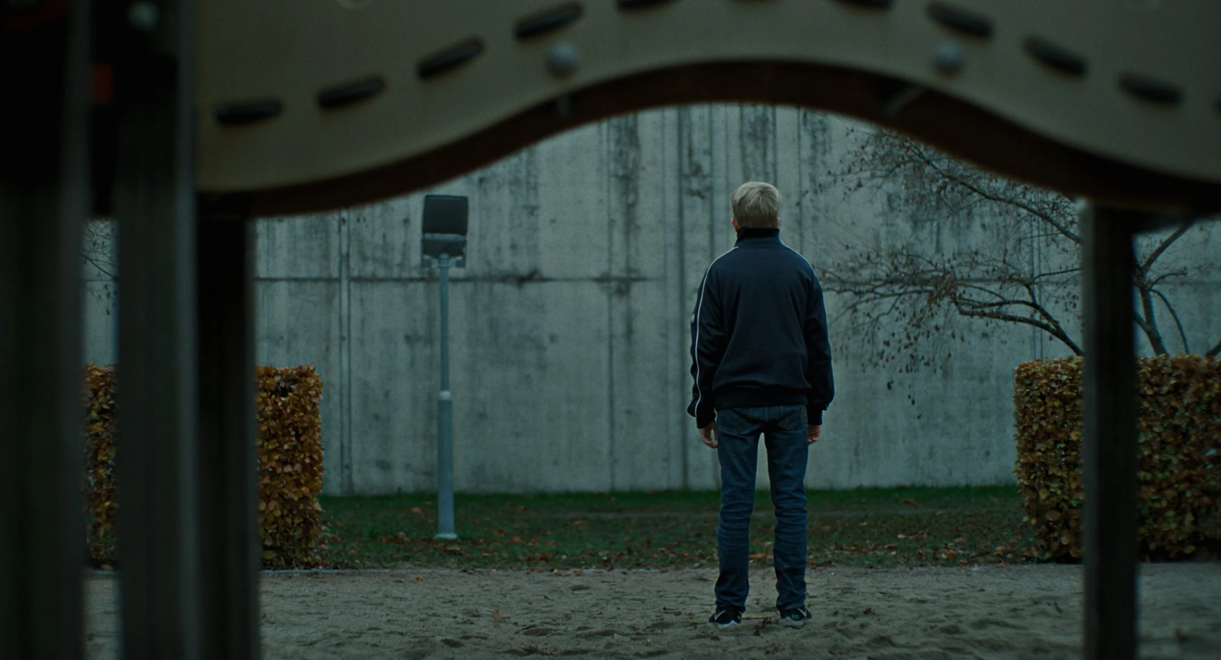 A man in a black jacket and jeans is standing alone on a dirt ground, facing a concrete wall with a basketball hoop nearby, seen from behind, surrounded by some bushes, in an outdoor setting.