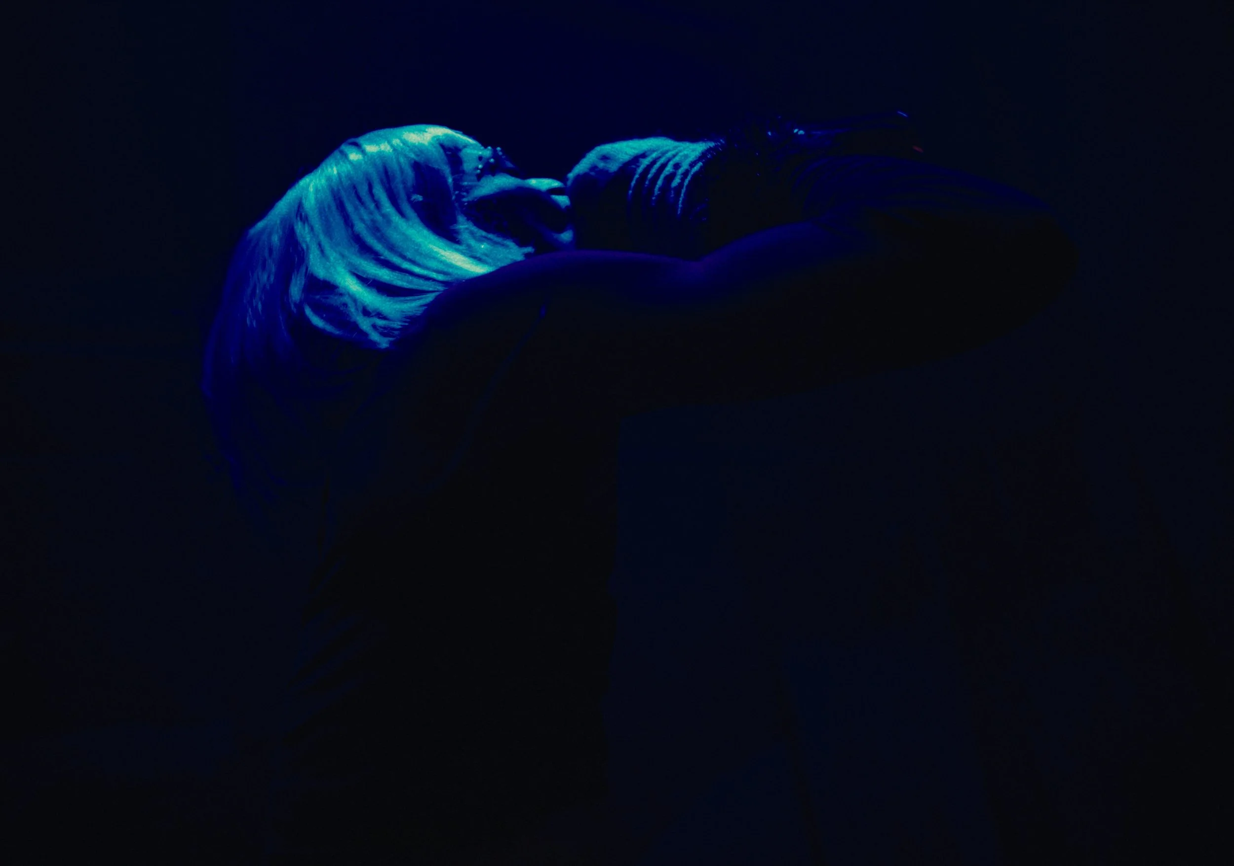 Person with blue neon light makeup and hair, posing with their head tilted back, in dark setting.