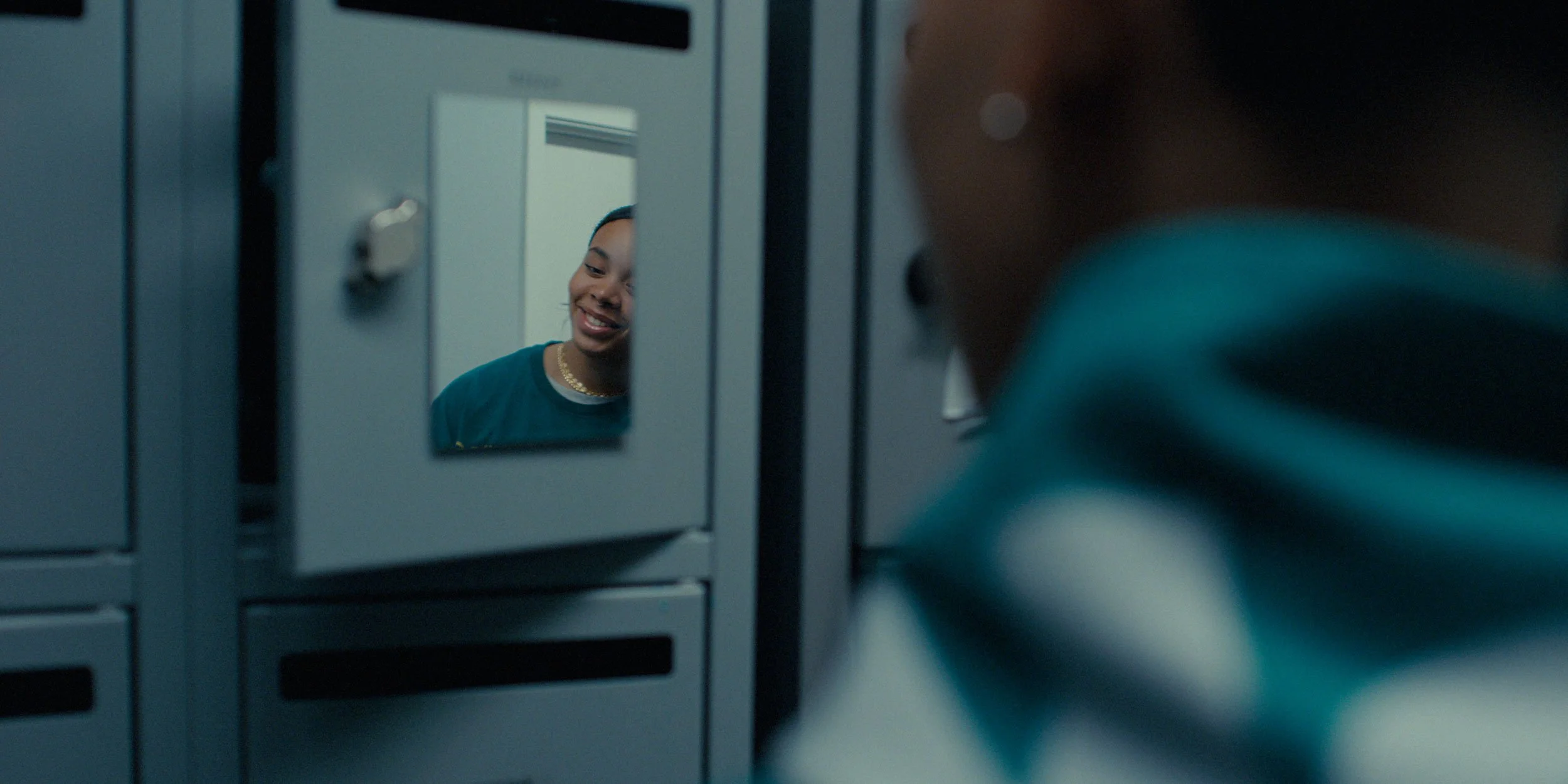 A person looking at their reflection in a small locker mirror, smiling, with lockers visible around them.