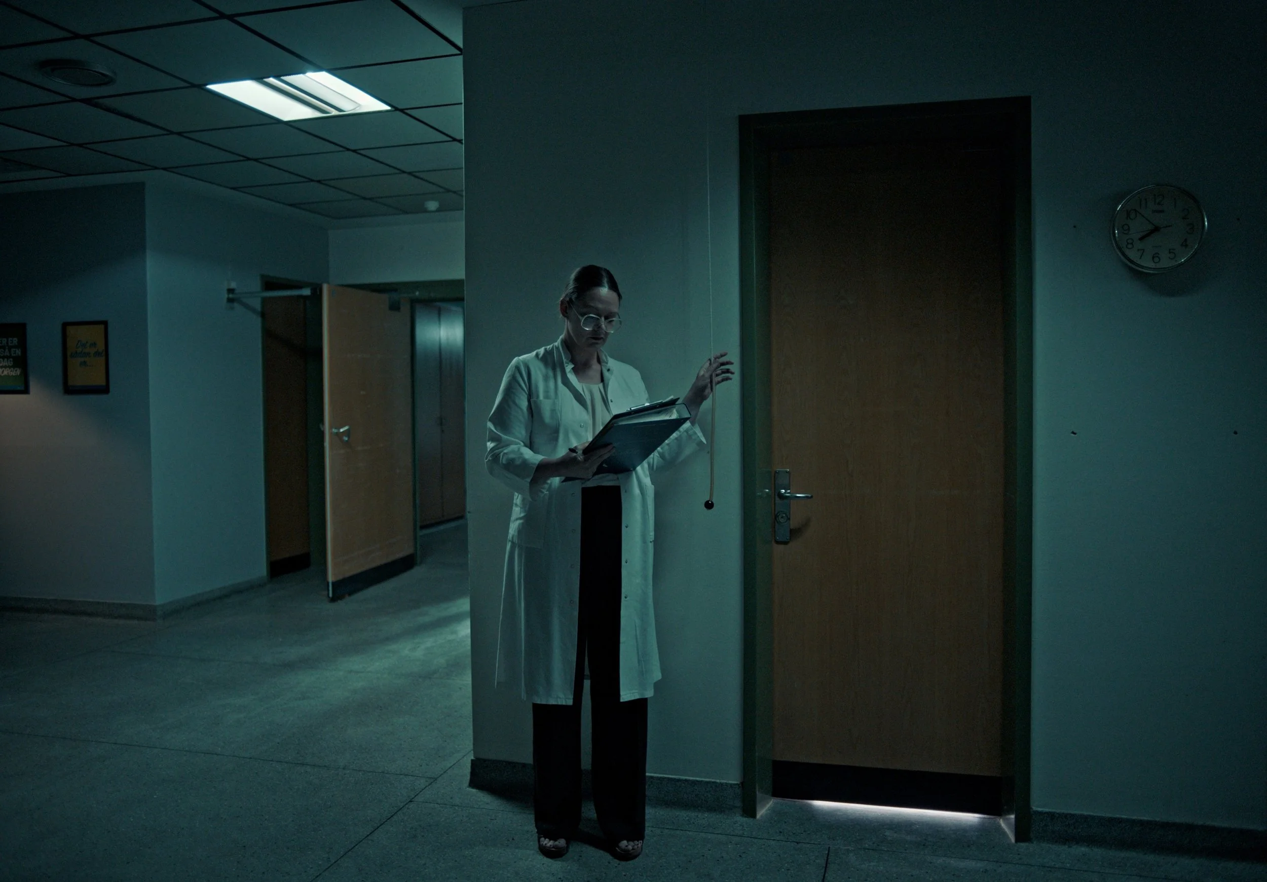 A woman in a white lab coat and glasses standing in a dimly lit medical or hospital corridor, reading a clipboard. There are lockers and a door nearby, and a clock on the wall showing around 9:15.