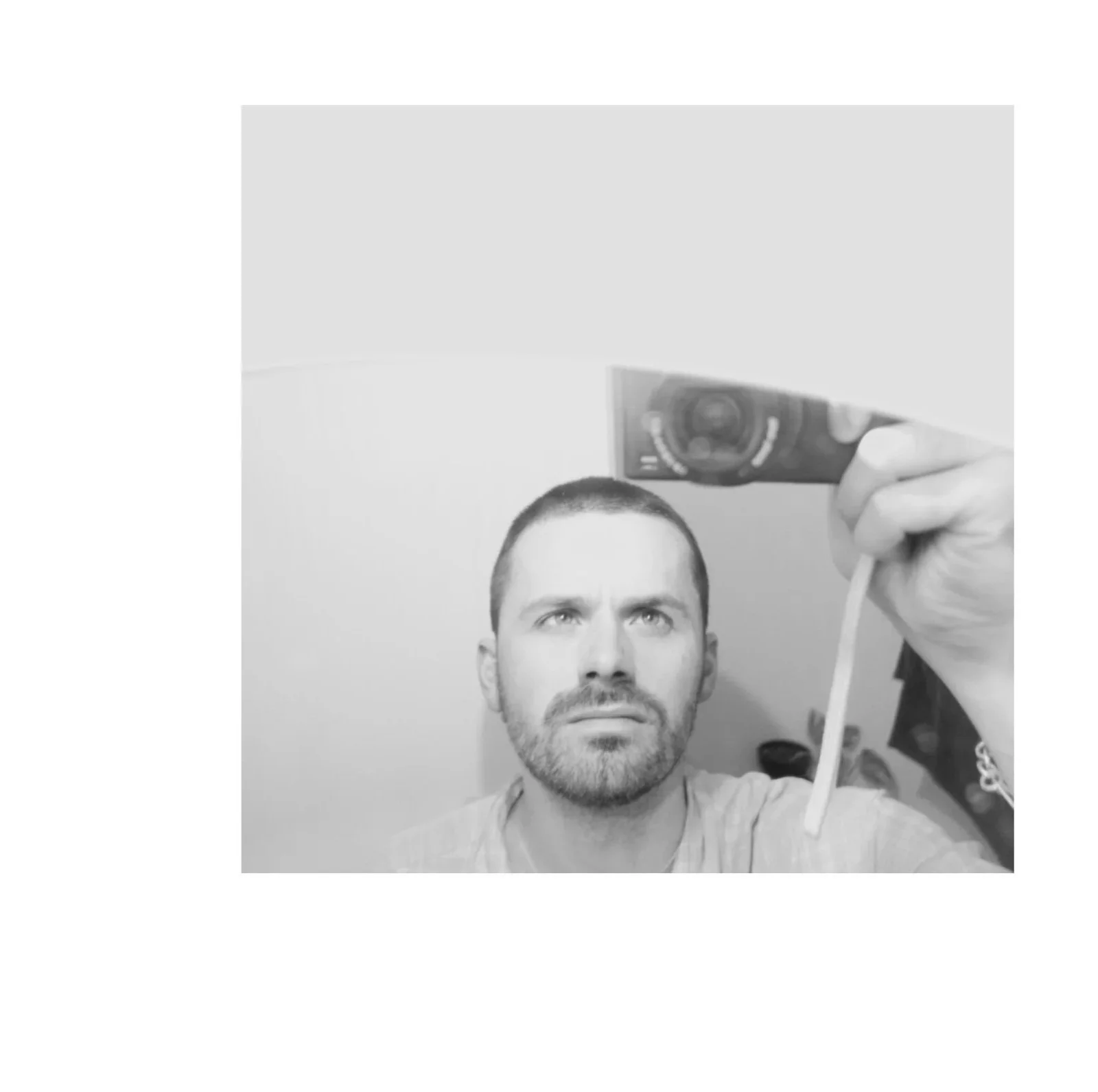 A black-and-white selfie of a man with short hair and a beard taking a photo with a small camera, with a serious expression, in front of a plain wall.