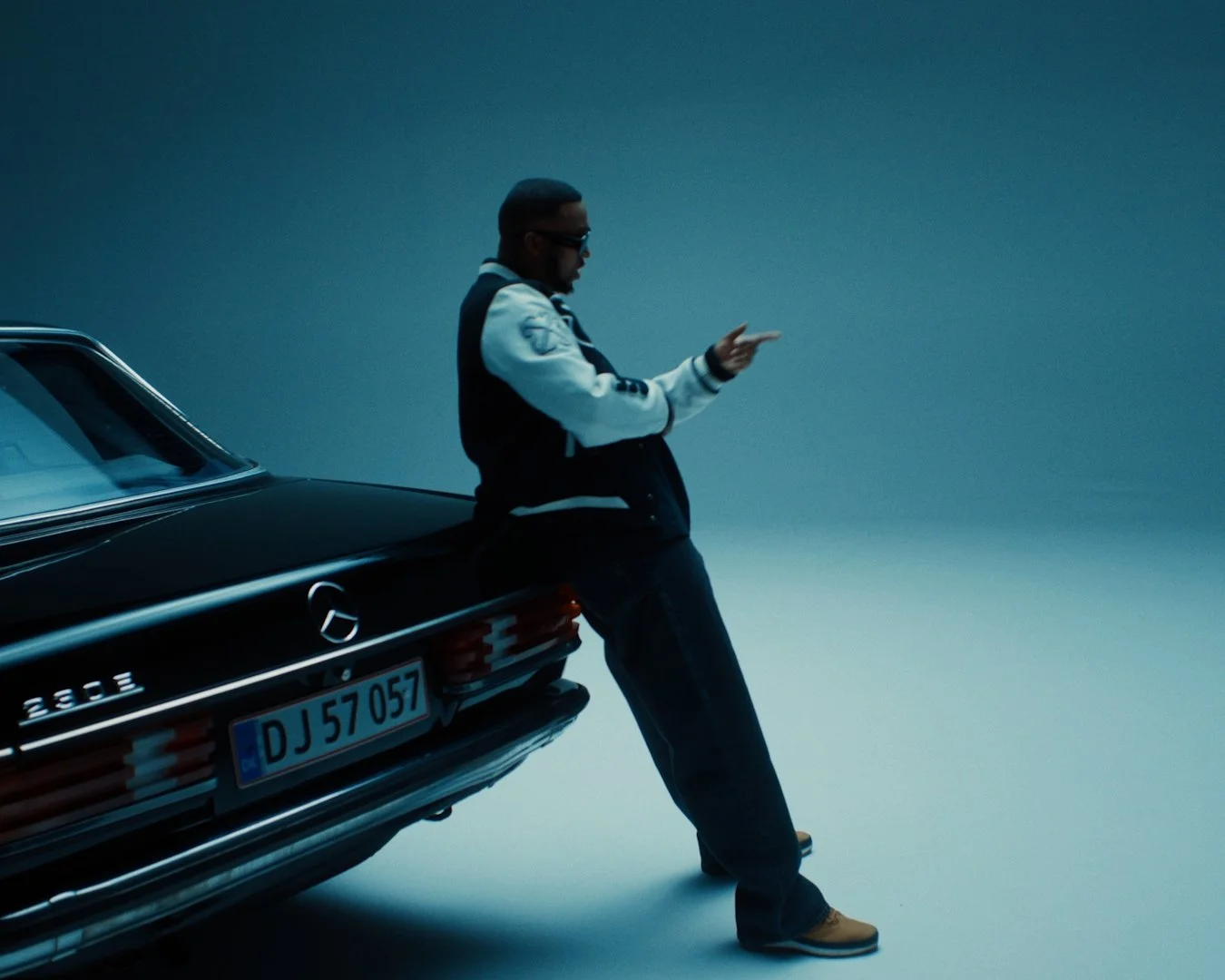 A man leaning against a vintage black Mercedes-Benz car, looking at his phone in a studio with backlighting
