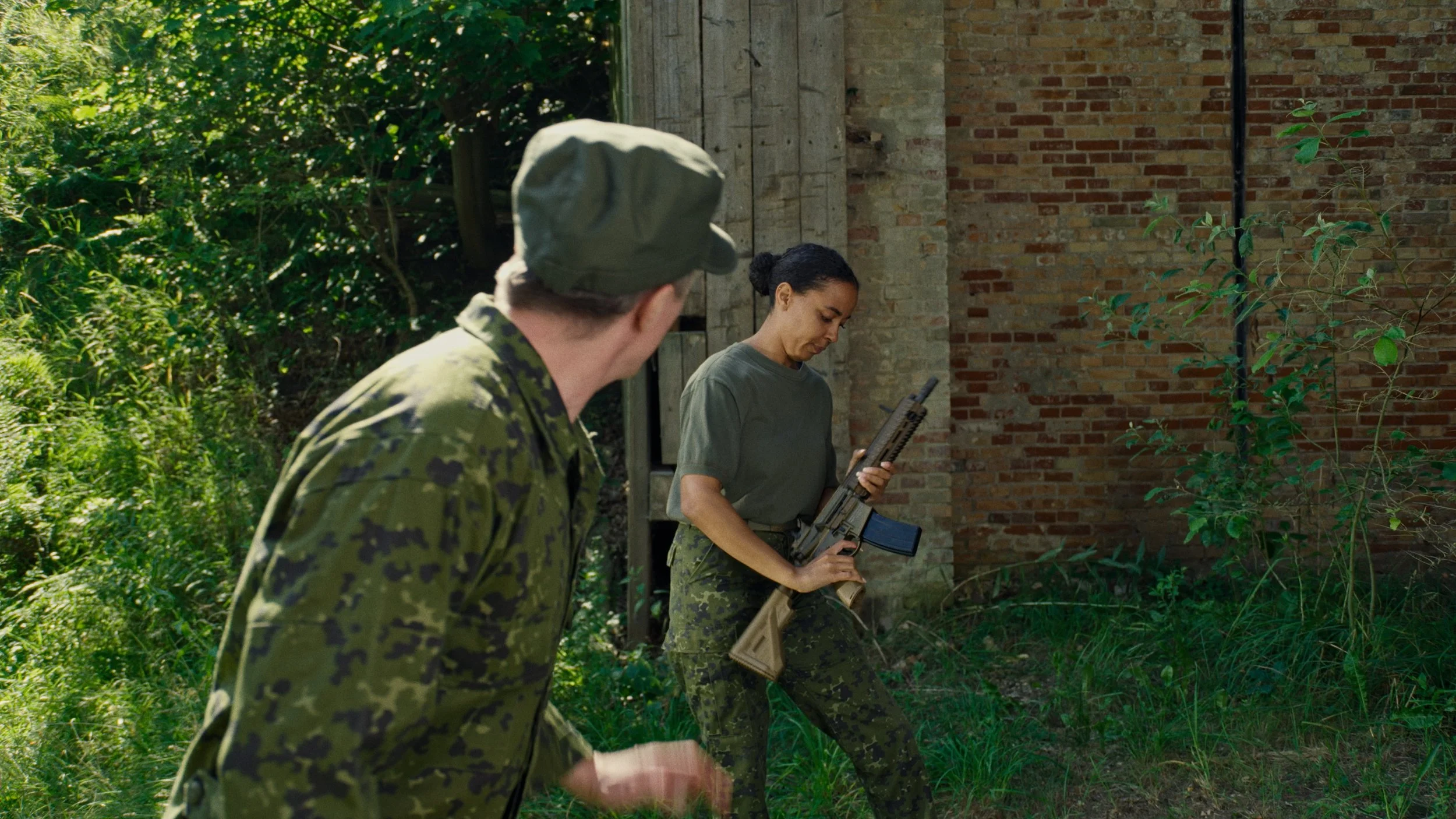 Two women in military camouflage uniforms, one holding a rifle, are outdoors near a brick building with greenery around.
