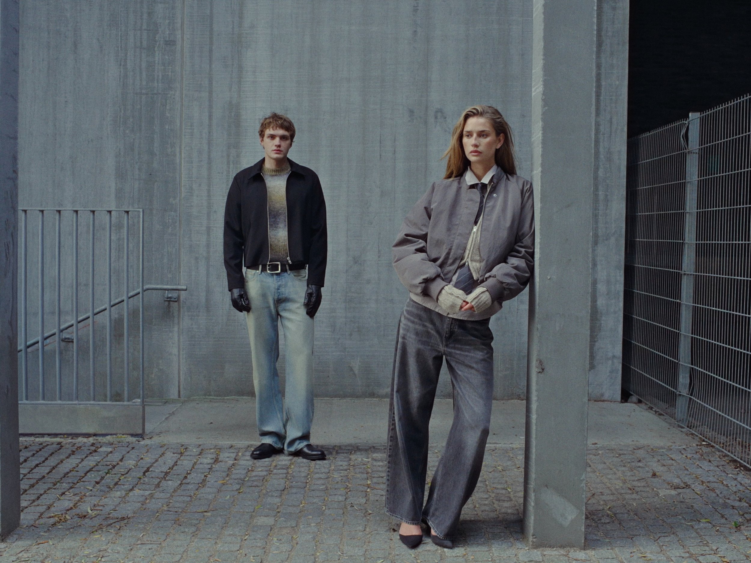 A young woman leaning against a concrete pole in front of a gray concrete wall, with a young man standing in the background on the cobblestone ground, both dressed in casual clothing.