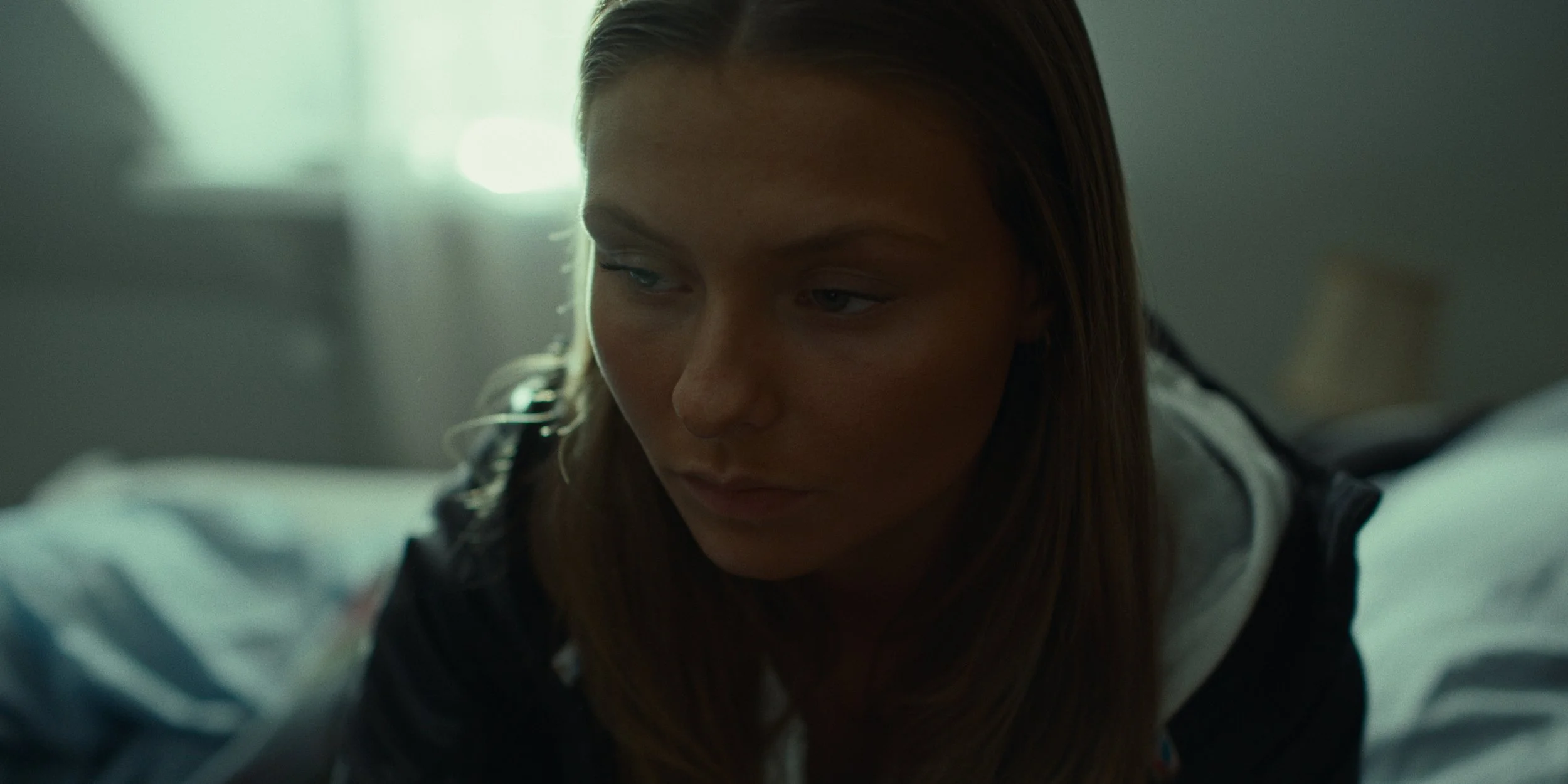A young woman with long brown hair and freckles resting on a bed, looking down thoughtfully in a softly lit room.