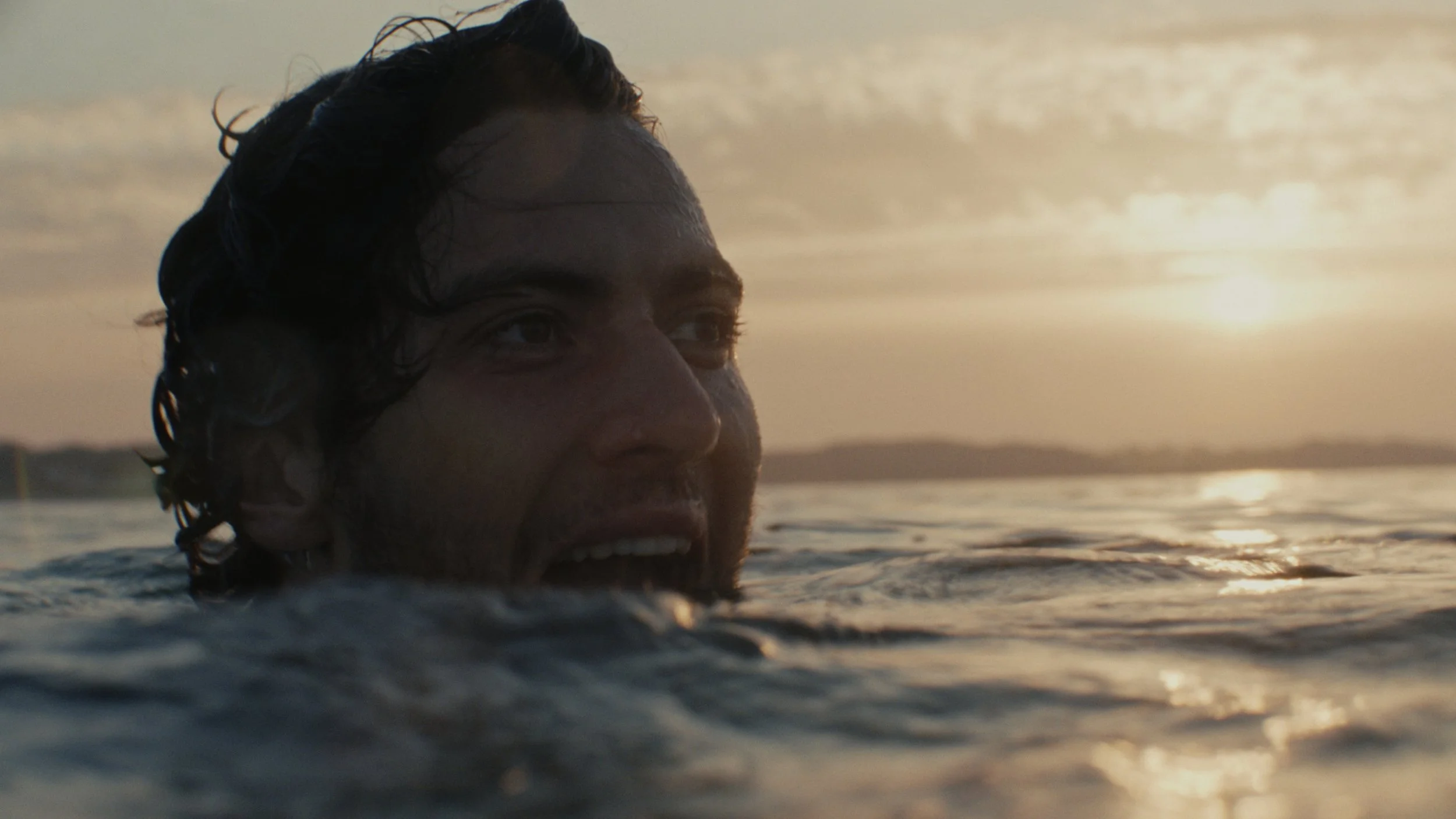 Close-up of a man swimming in a body of water with the sunset in the background, his face partially submerged and water splashing around him.