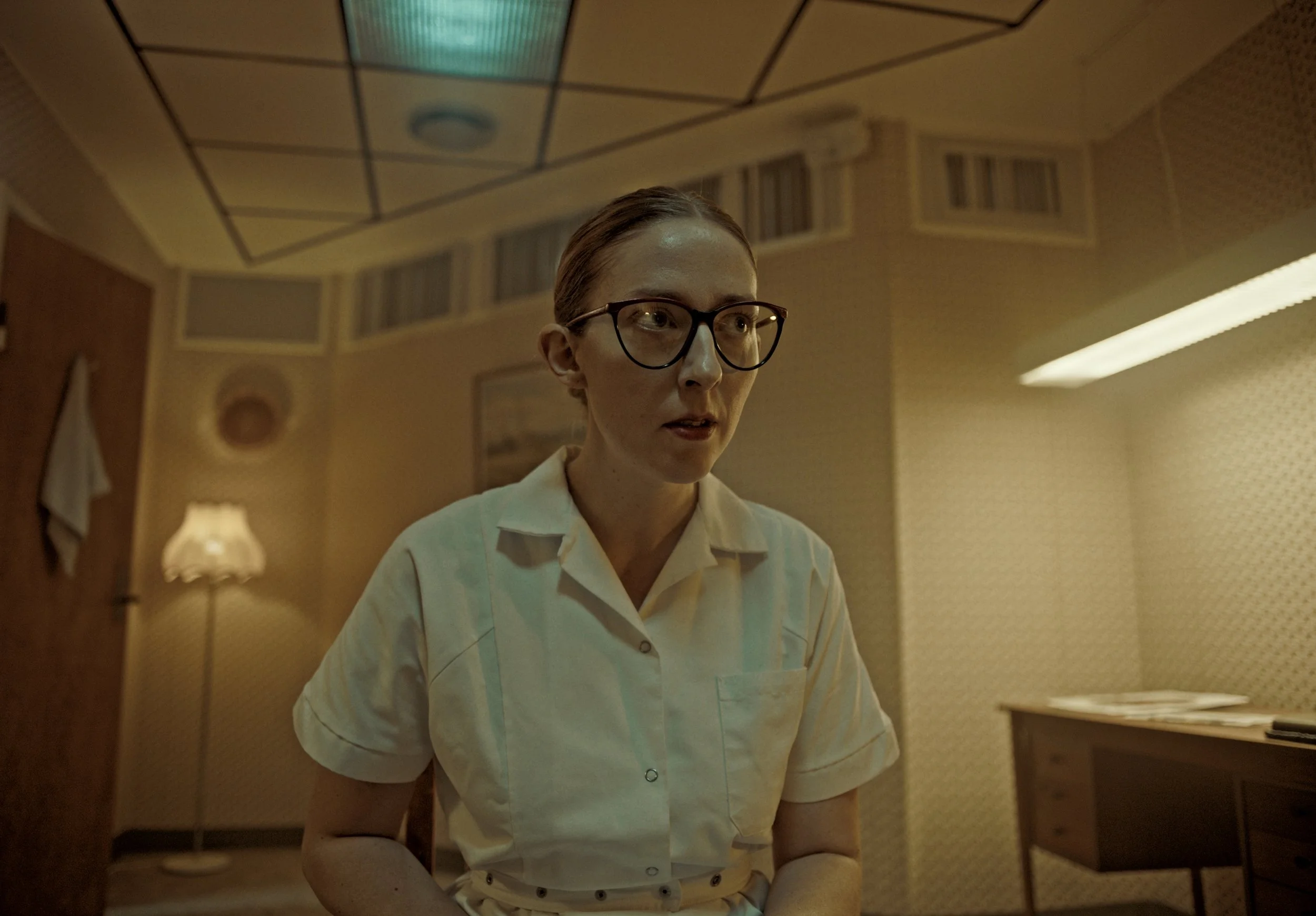 A woman with glasses and a beige uniform standing in a dimly lit room, possibly an office or a police station, with patterned wallpaper, a framed picture on the wall, a lamp, and a side table.
