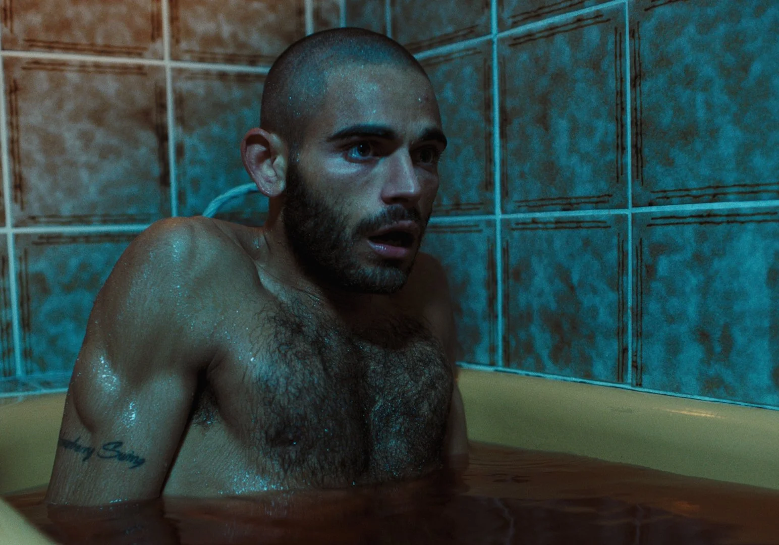 A shirtless man with a beard and tattoos on his arm sits in a yellow bathtub, with water up to his chest, in a tiled bathroom. He appears surprised or startled.