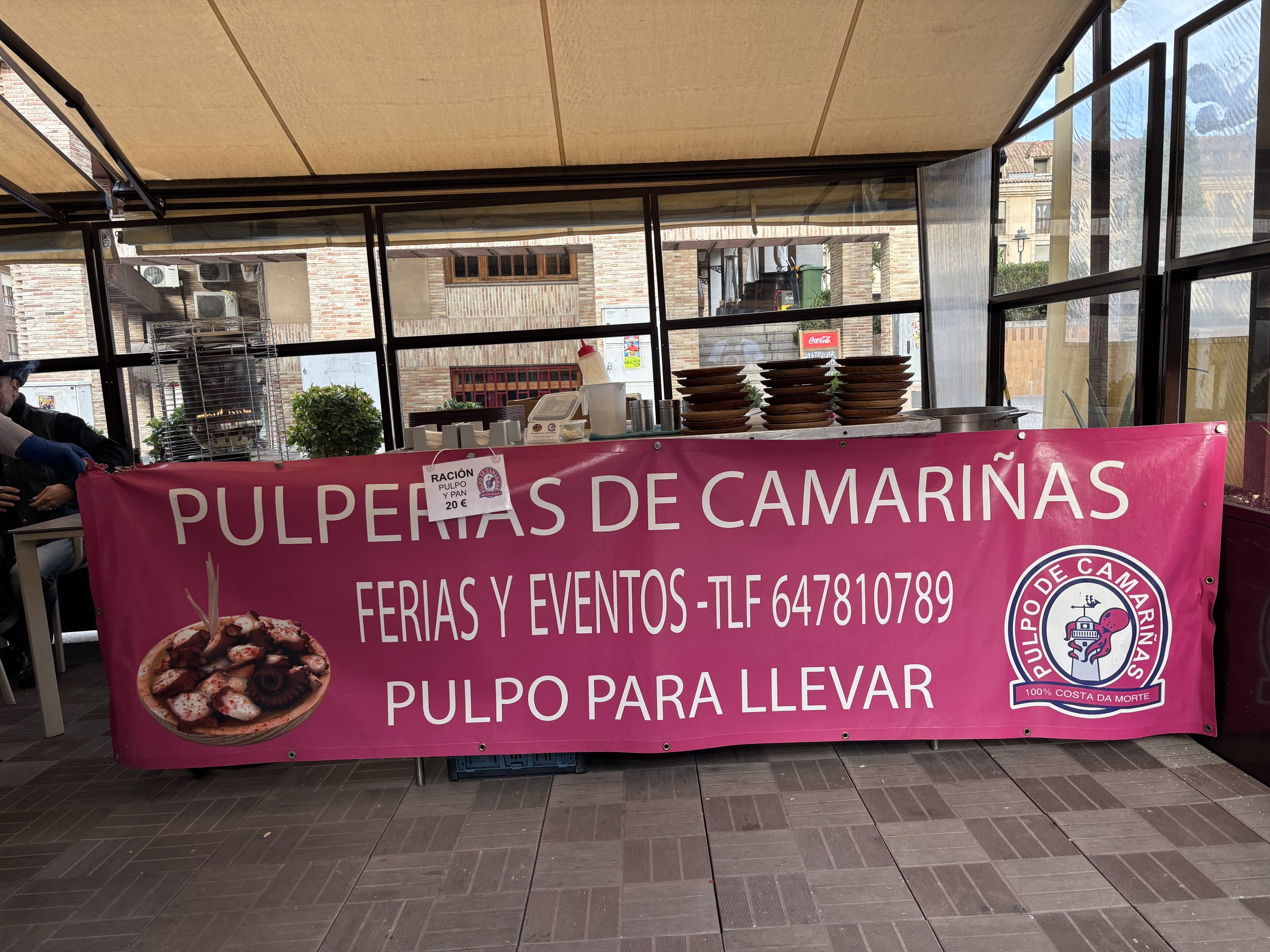 Cartel rosa que anuncia pulpo de camariñas para ferias y eventos, con número de teléfono. En la mesa, platos y algunos ingredientes para preparar el pulpo.