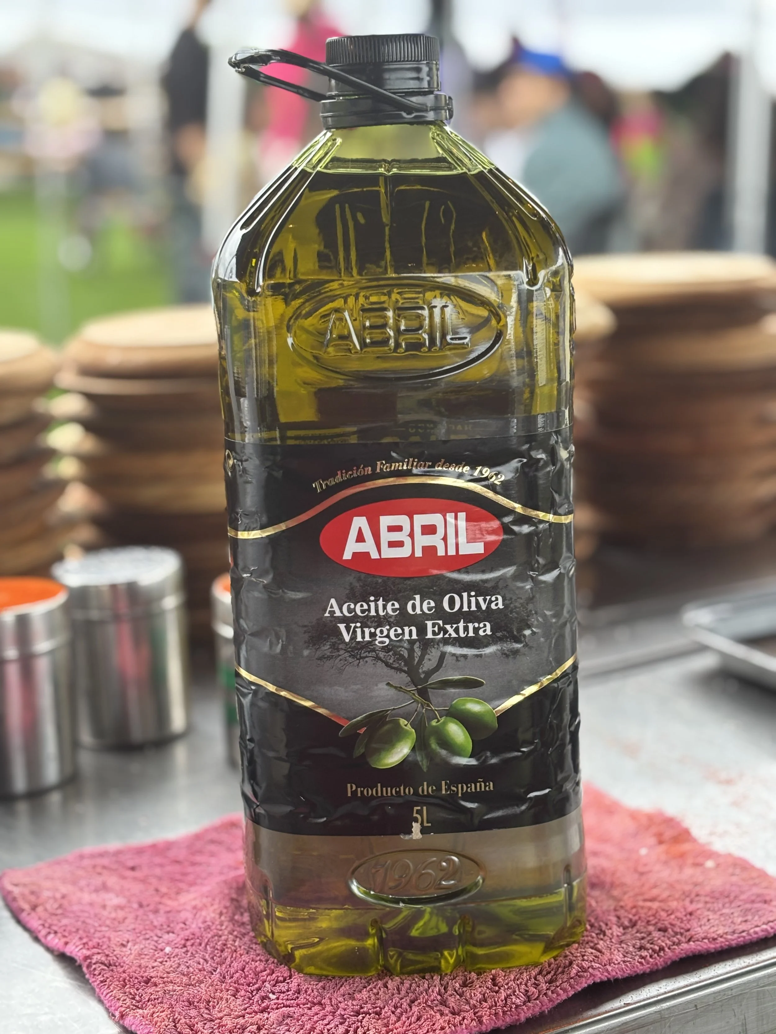 Botella de 5 litros de aceite de oliva virgen extra marca Abril, con etiqueta negra y rojo, sobre una toalla rosada.