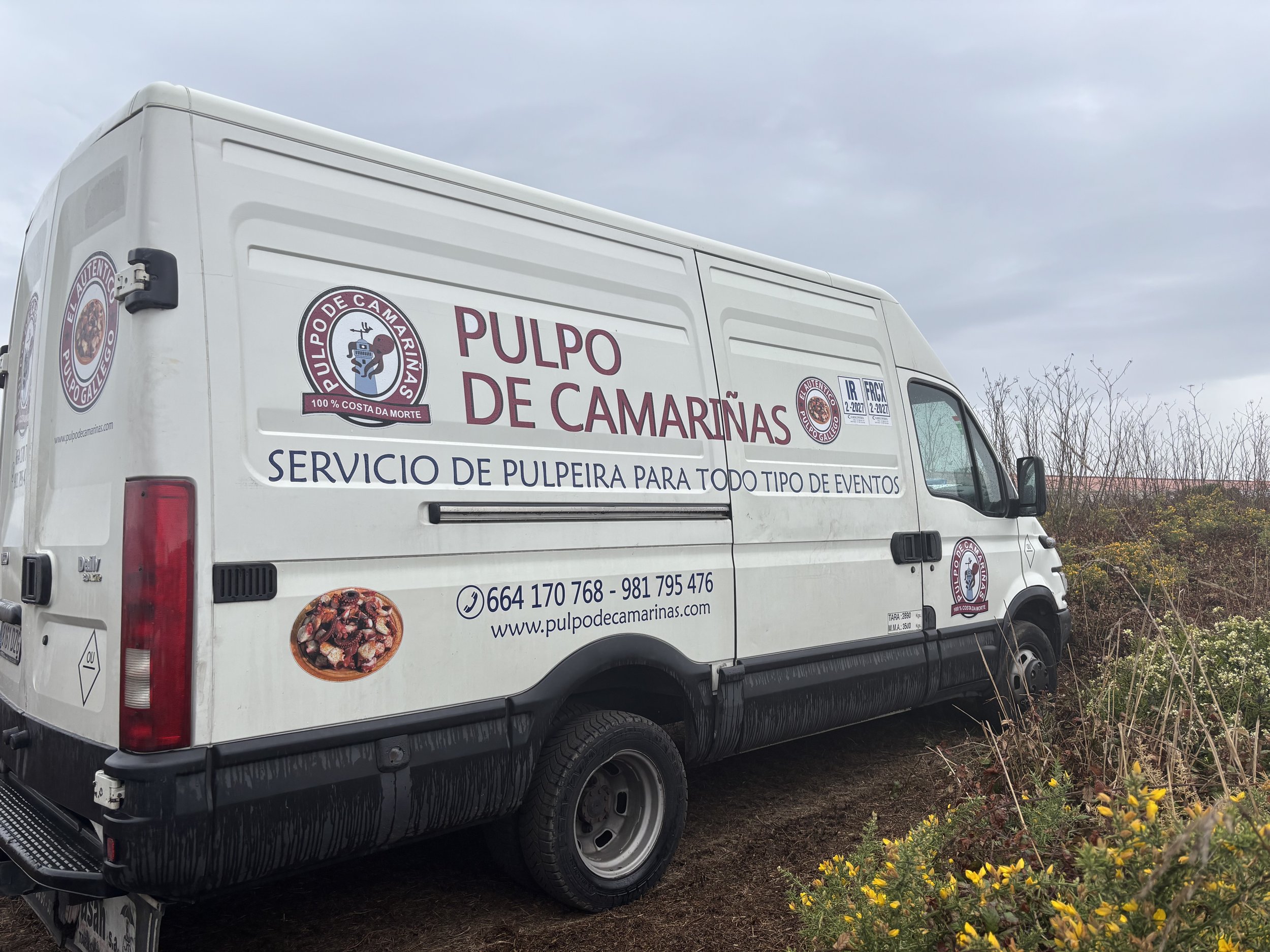 Camioneta blanca con etiquetas y logotipos que promocionan pulpo de camariñas, un servicio de pulpo para eventos, estacionada en un área con arbustos y flores en un día nublado.