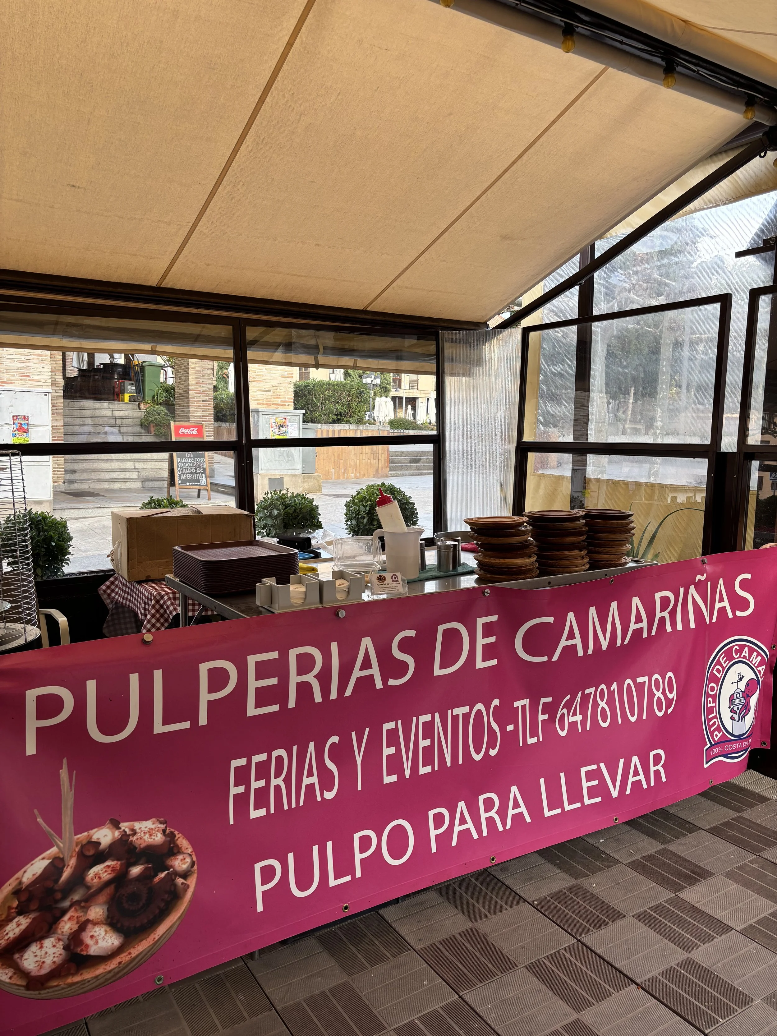 Carpa con mesa de pulpería de camarinas, platos, utensilios y un cartel rosa con información de contacto y servicios de la venta y eventos de pulpo para llevar.