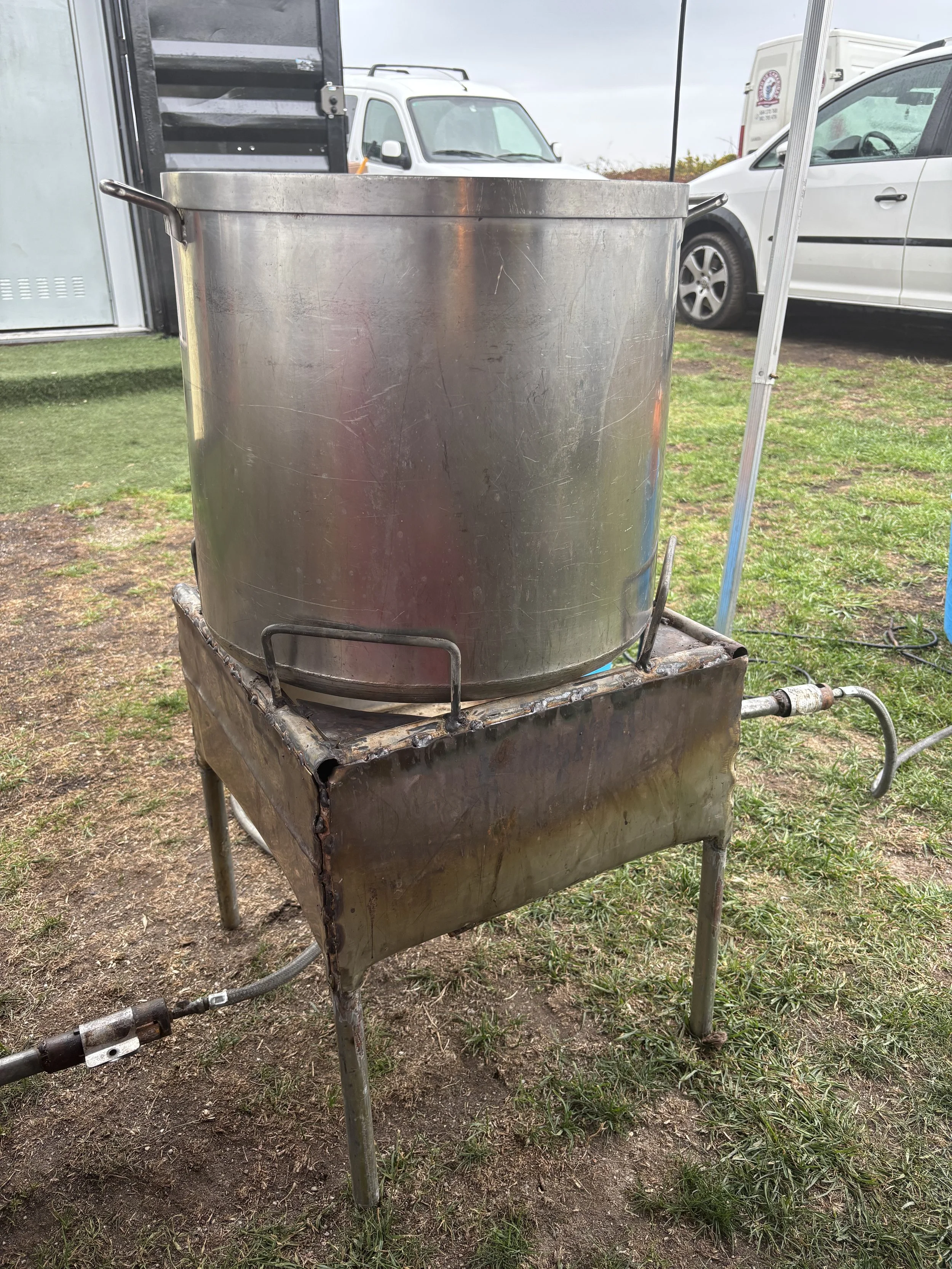 Olla grande de metal sobre una parrilla de metal con ruedas, en un ambiente al aire libre.