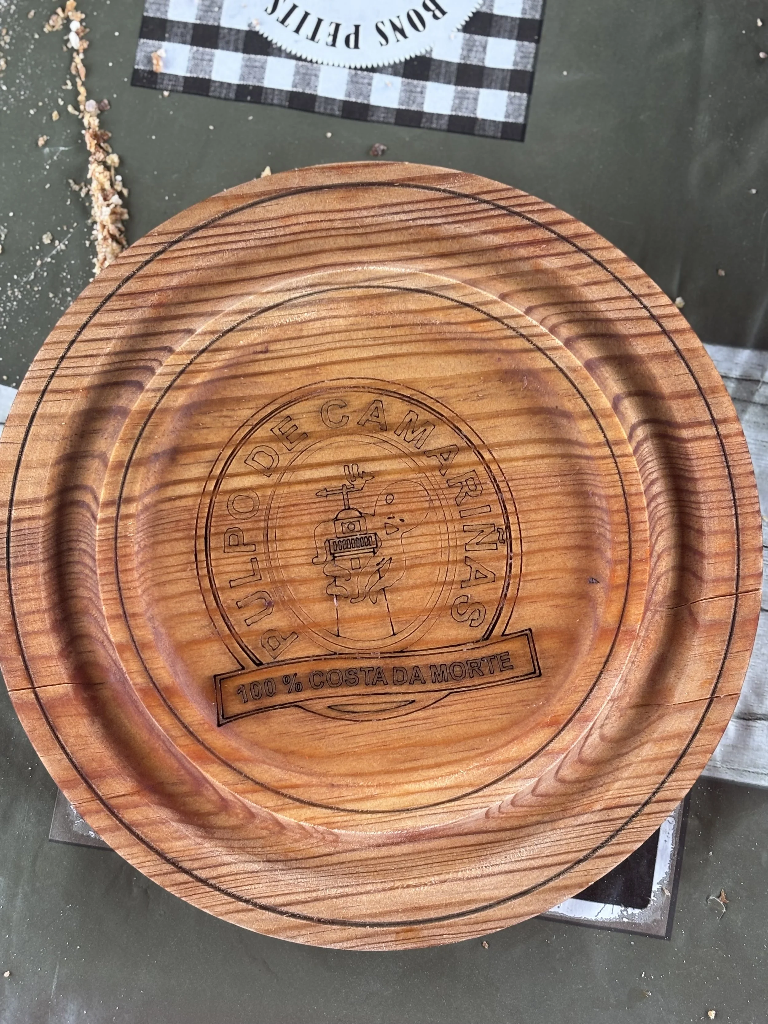 Plato de madera con grabado que dice 'Pulpo de Camarinas' y una imagen de un pulpo con una cámara zapando, con la frase '100% Costada Morte' en la parte inferior.